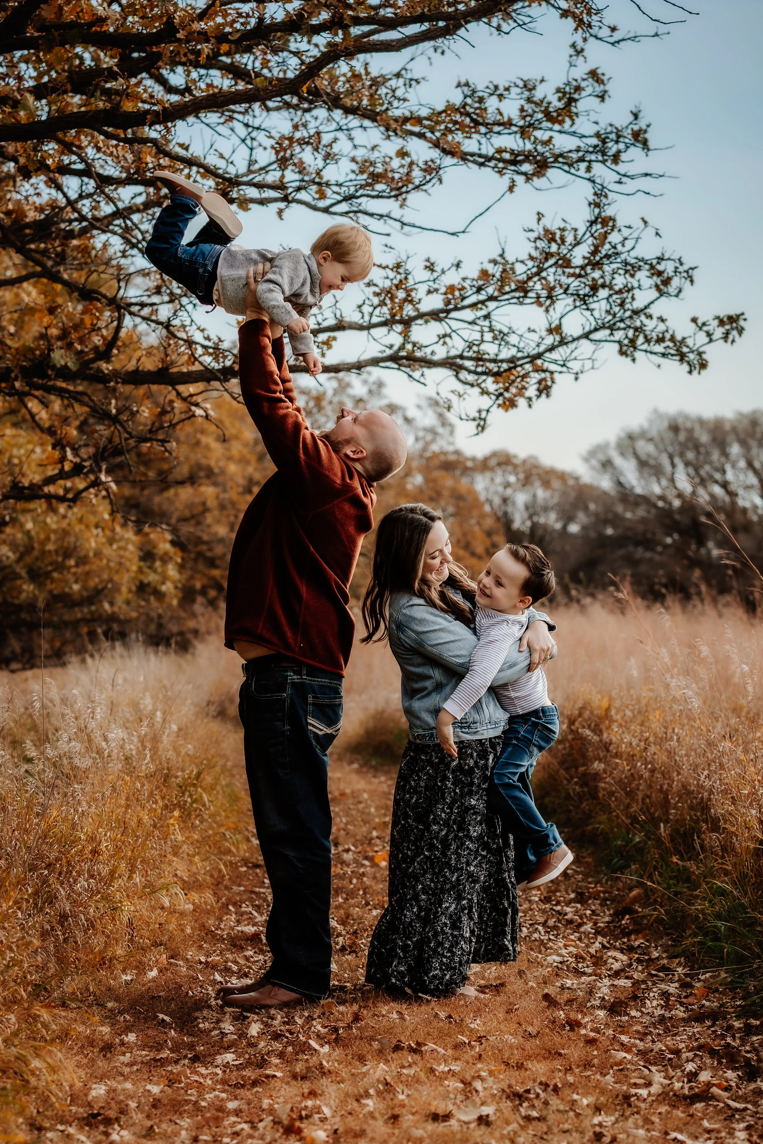 Family of four playing outdoors on a fall day with orange-tinted grass and trees in the background. The father holds a young boy up in the air, while the mother and an older boy embrace and smile in Sioux Falls, South Dakota at Good Earth State Park.