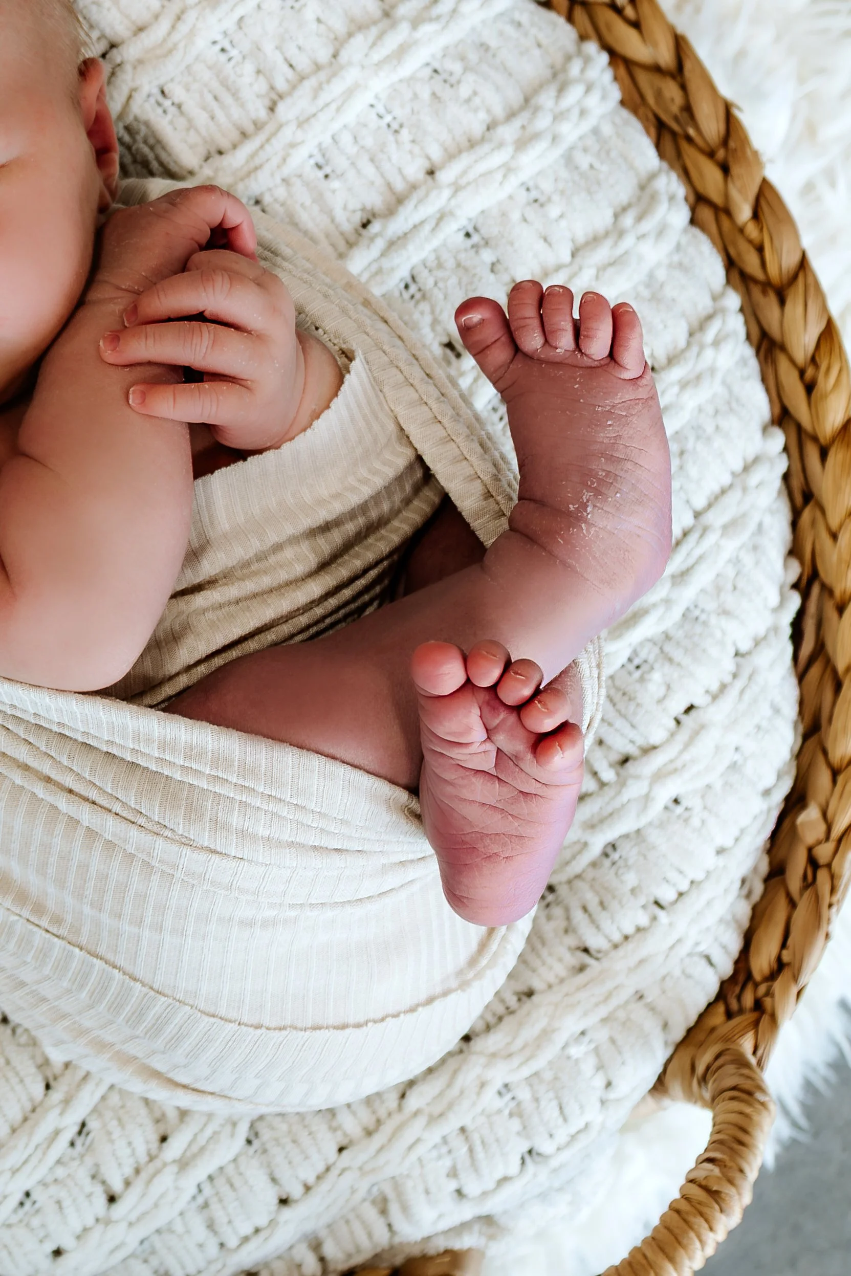 Close-up of a newborn baby lying in a woven bassinet, wrapped in a cream-colored blanket, with tiny feet and hands visible, some skin peeling and peeling on the feet at The White Space Studio in Sioux Falls, South Dakota.