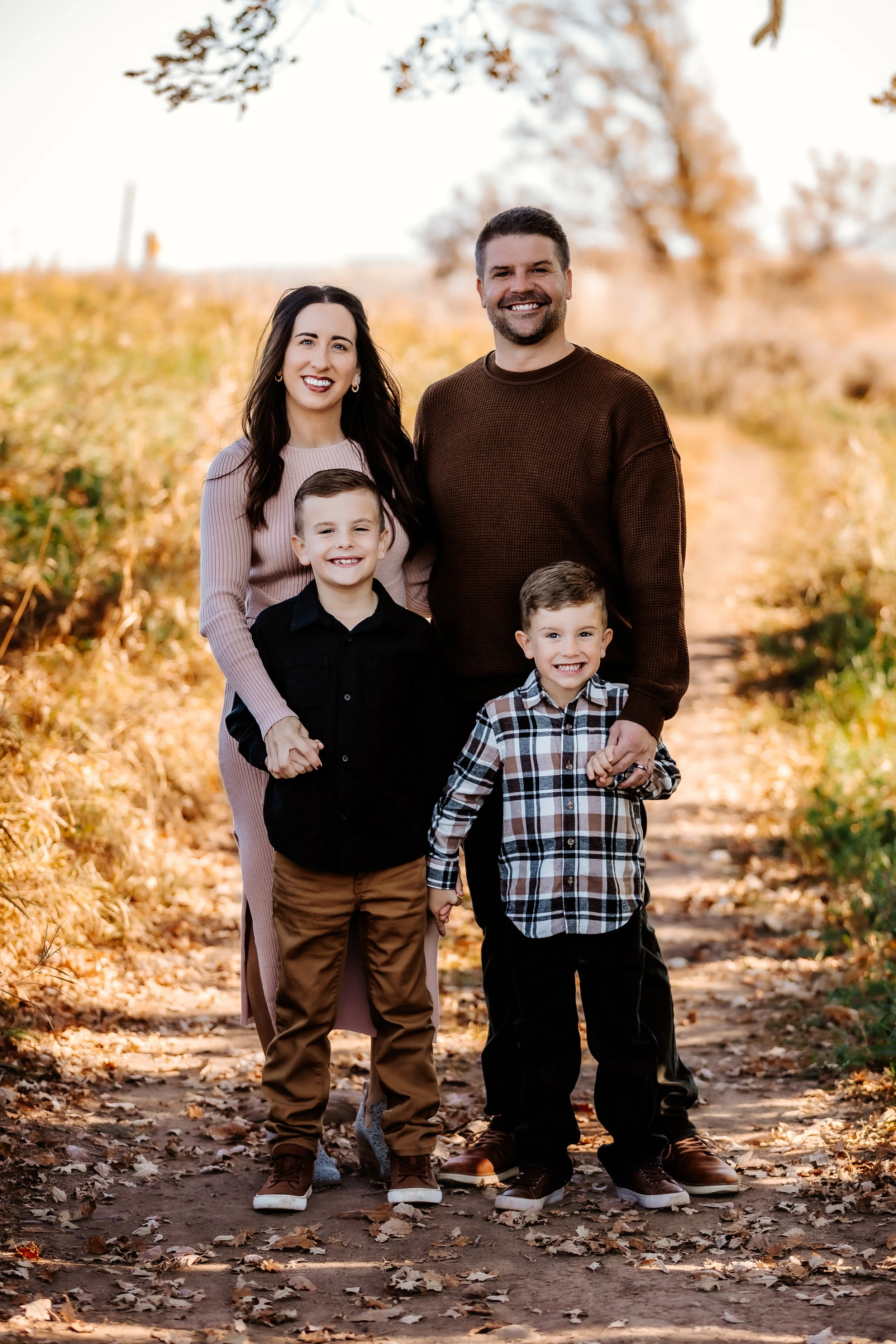 A family of four standing on a leaf-covered trail in a park during autumn. The mother, father, and two young boys are smiling at the camera with trees in the background in Sioux Falls, South Dakota at Good Earth State Park.