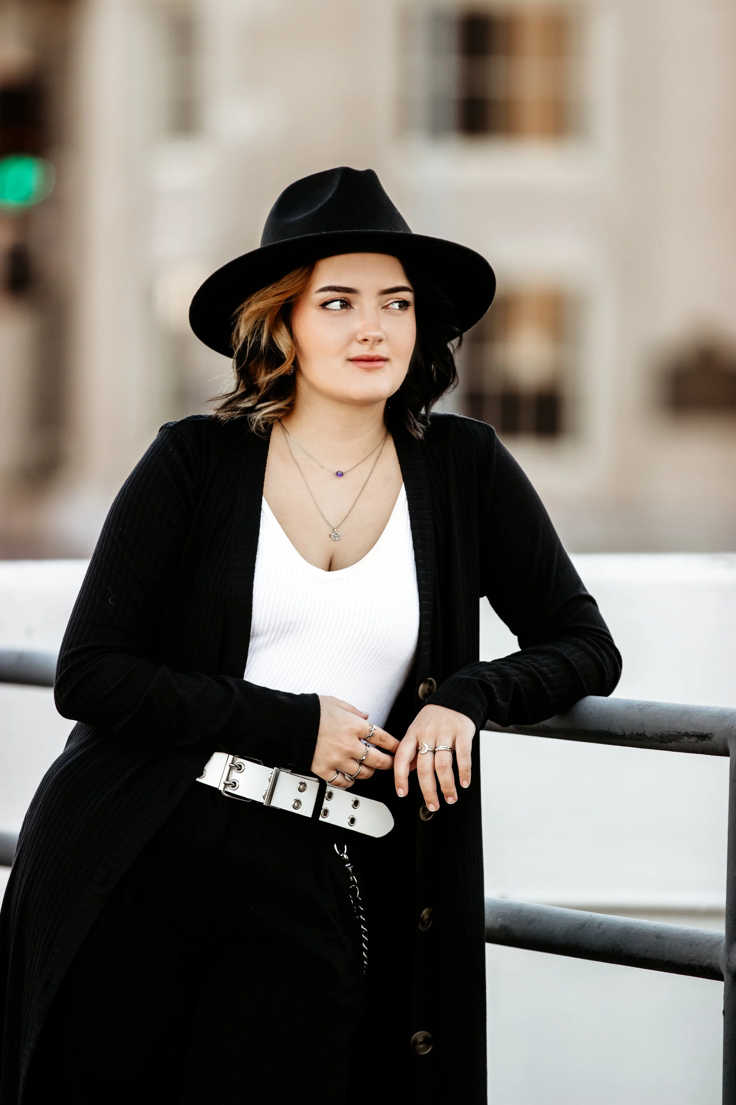 A young woman with brown wavy hair wearing a black wide-brimmed hat, black cardigan, white top, black pants, and jewelry, standing outdoors near a metal railing with blurred buildings in the background in Downtown Sioux Falls, South Dakota.