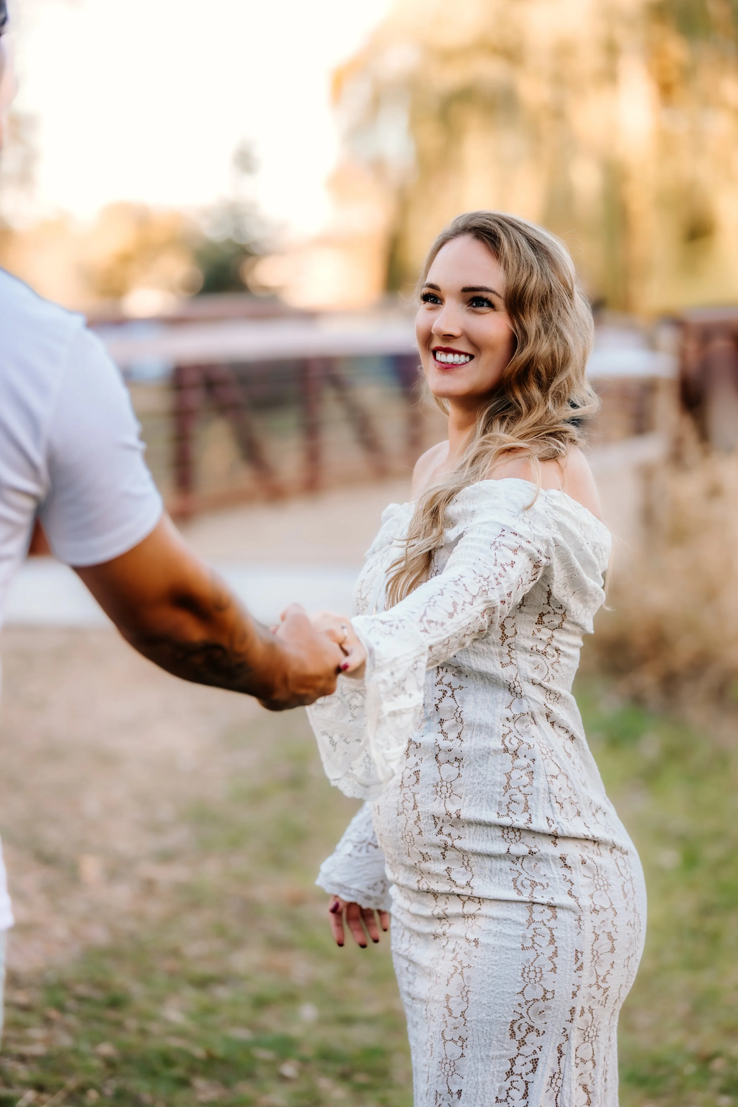 A smiling woman in a white lace dress holding hands with a man outdoors during daylight, with trees and a wooden fence in the background at Mary Jo Wegner Arboretum in Sioux Falls, South Dakota.
