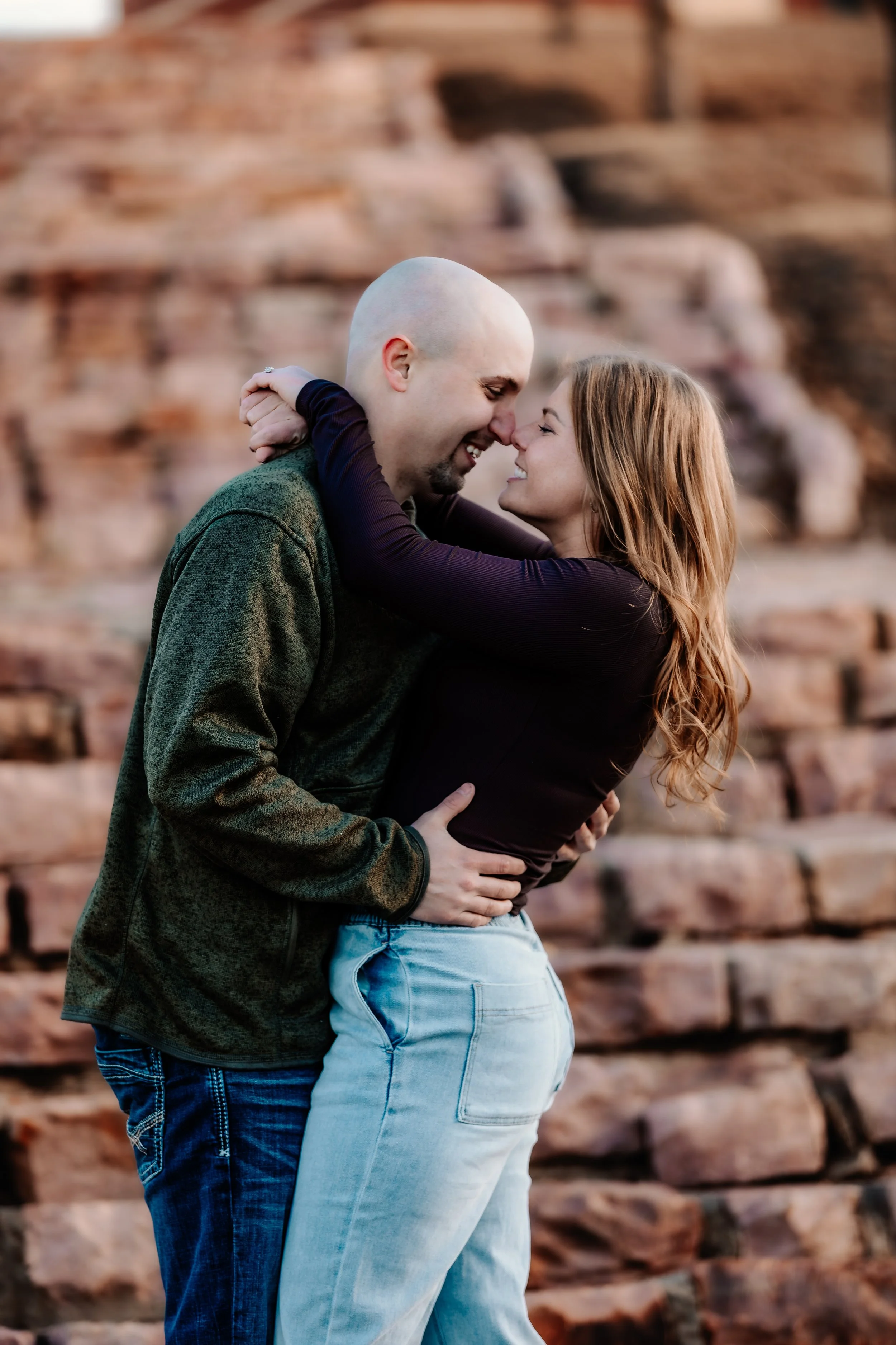 A couple embracing and smiling, touching foreheads outdoors with a rocky background, during sunset at Terrace Park in Sioux Falls, South Dakota.