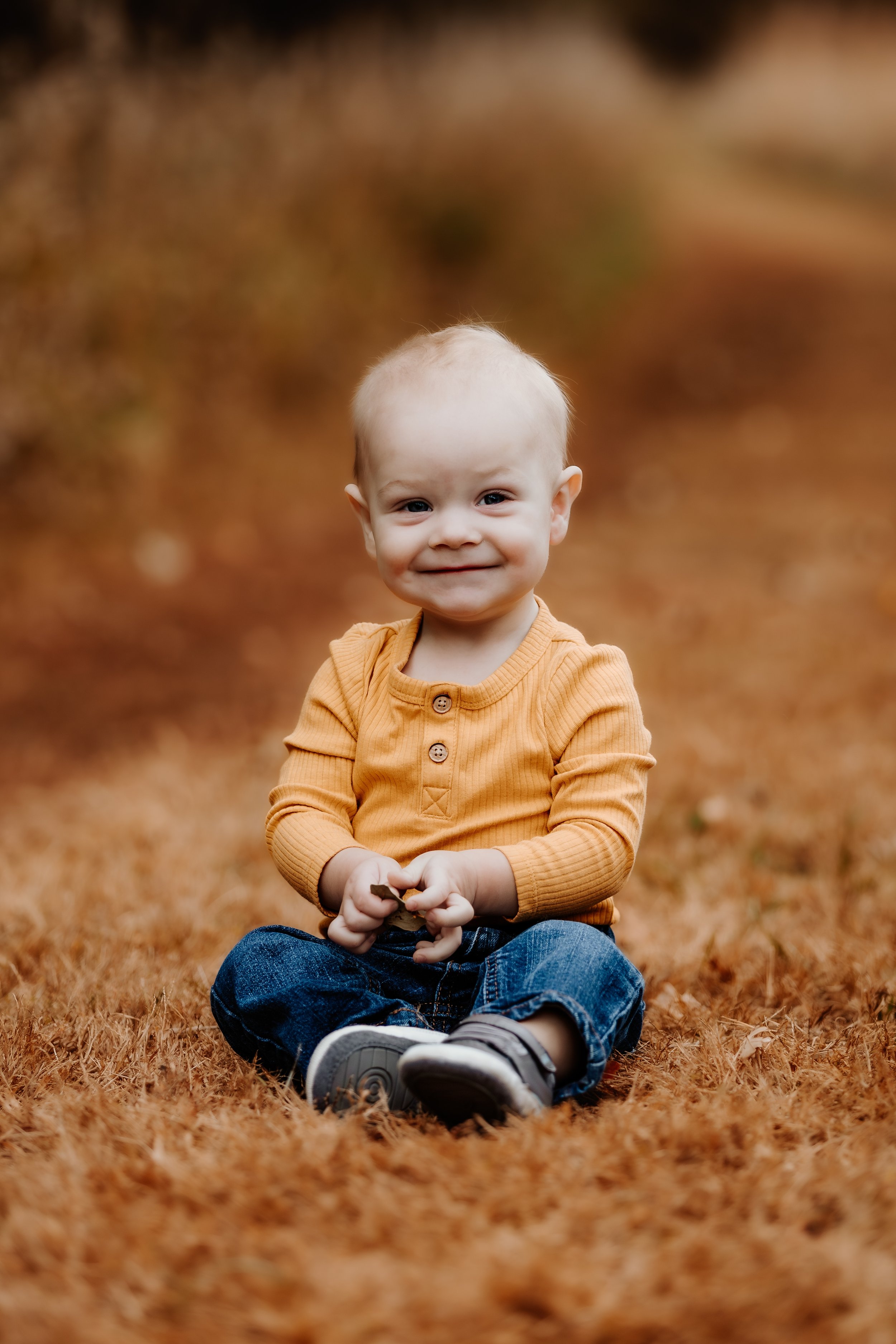 A young child sitting on a grassy area with brown foliage in the background, wearing an orange long-sleeve shirt and blue jeans, smiling and holding a small object in their hands in Sioux Falls, South Dakota at Good Earth State Park.