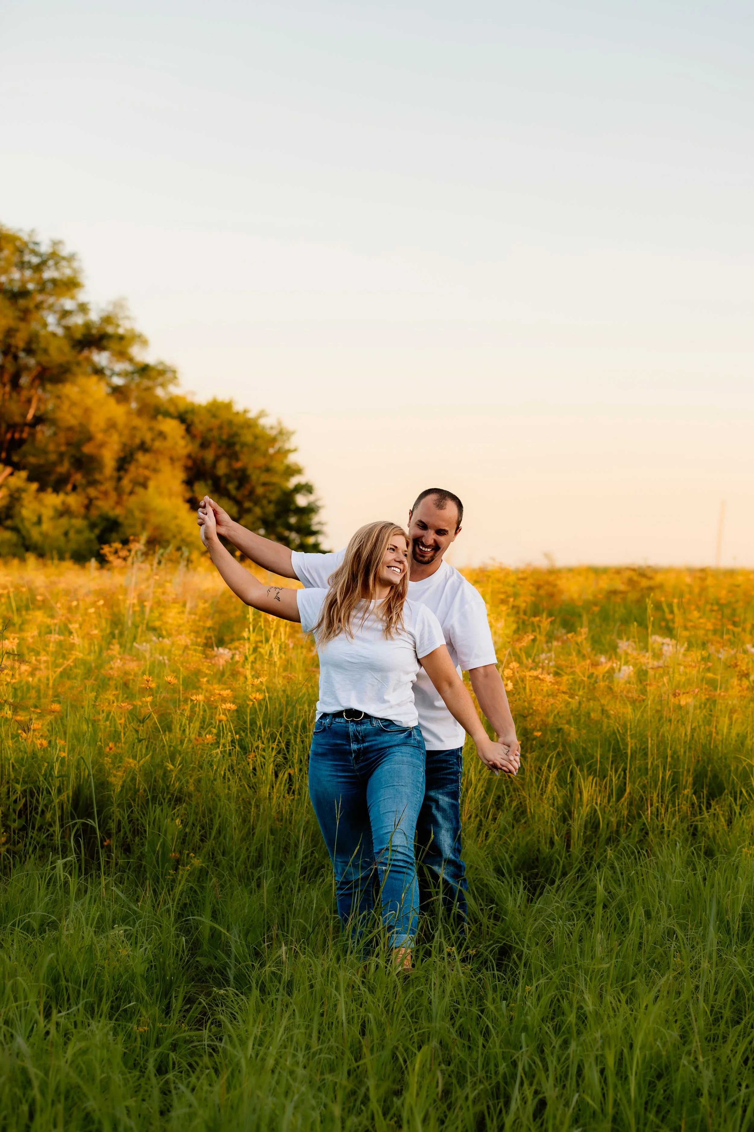 A happy couple holding hands and dancing in a field of tall grass and yellow wildflowers at sunset, with trees in the background at Good Earth State Park in Sioux Falls, South Dakota.