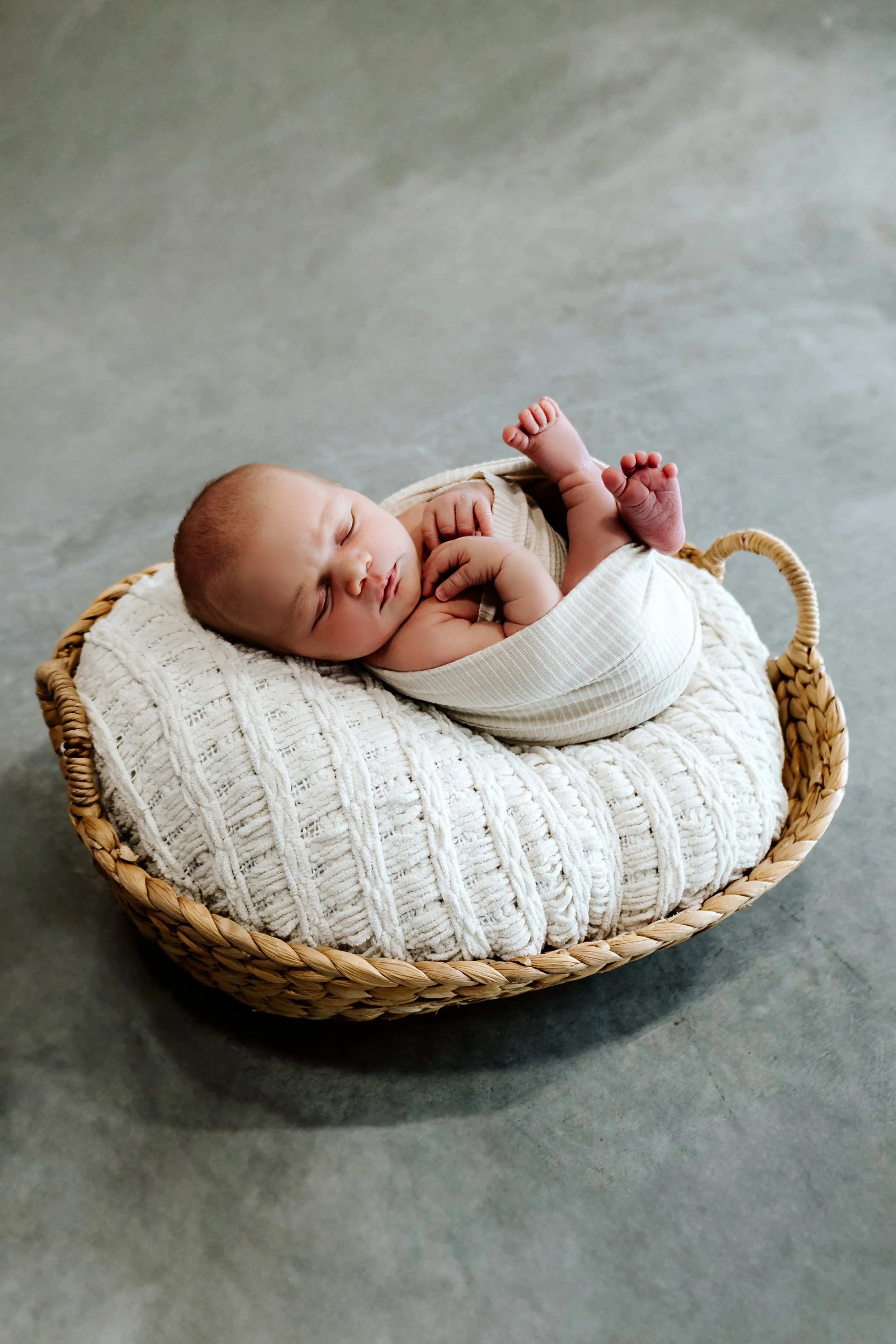 A newborn baby sleeping in a woven basket on a soft, textured blanket on a concrete floor at The White Space Studio in Sioux Falls, South Dakota.
