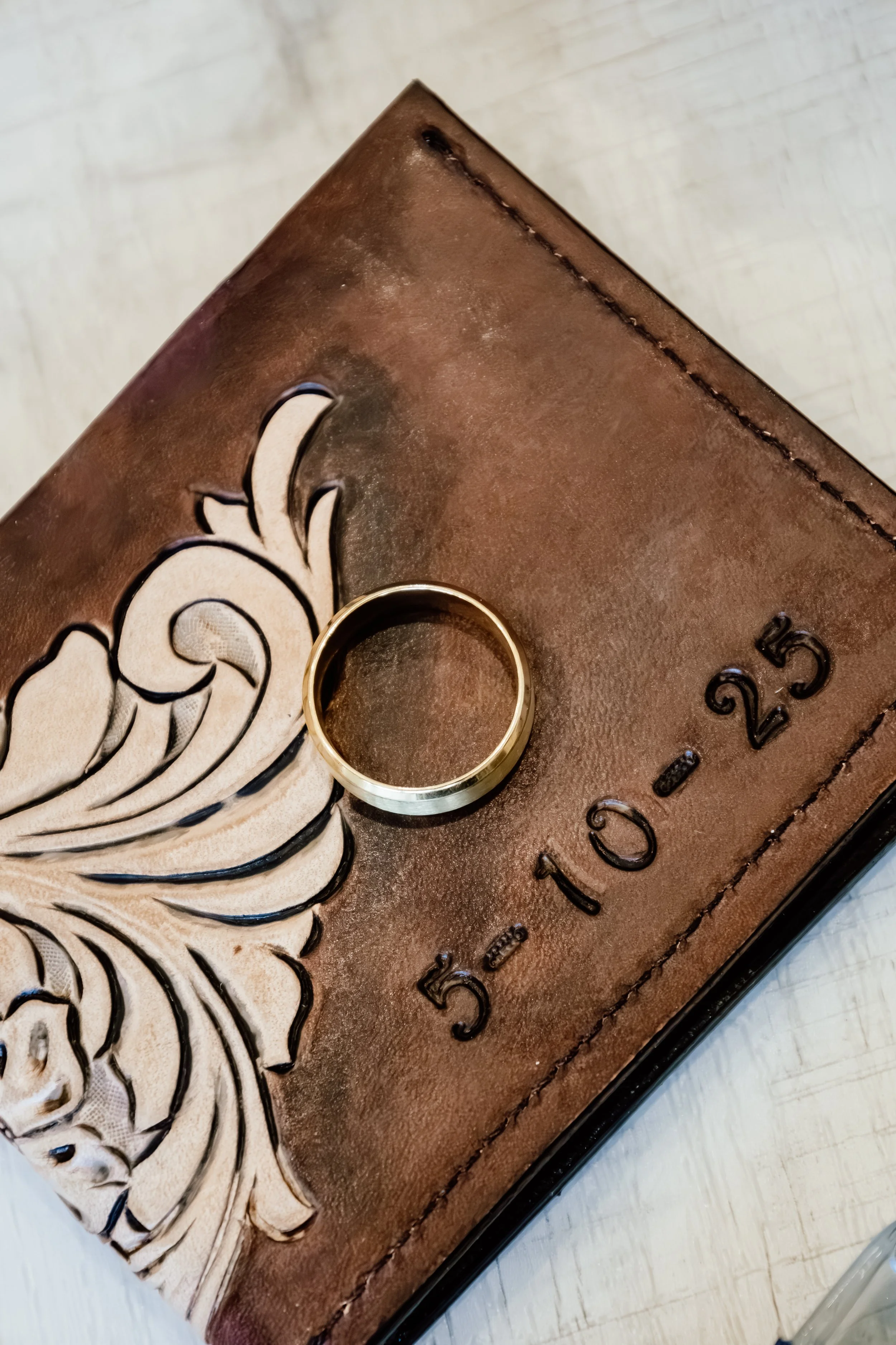 A gold wedding band ring resting on a brown leather wallet with a floral design, and the date '5-10-23' embossed on it at Laurel Ridge in Sioux Falls SD.