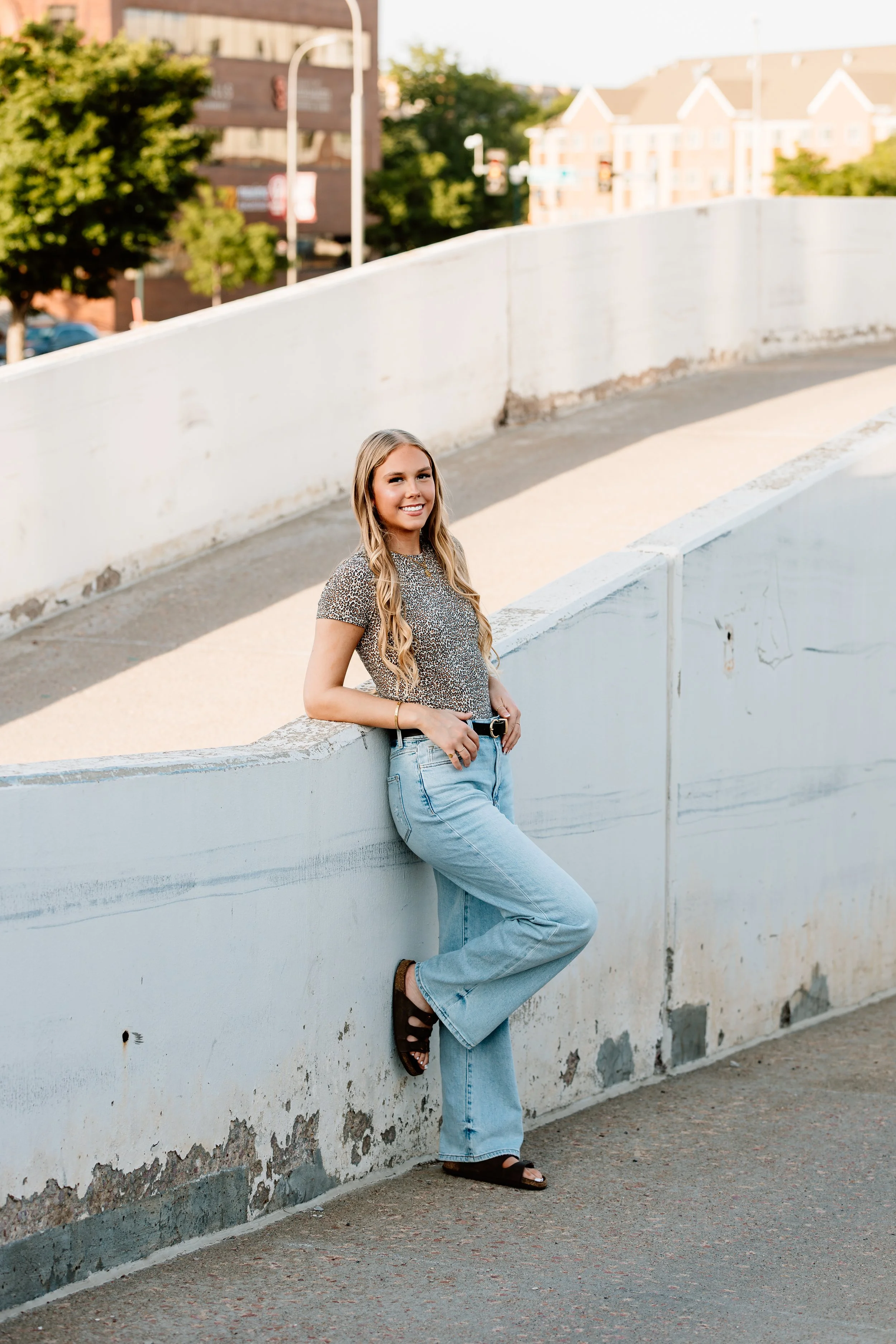 A young woman with long blonde hair styled in loose waves, smiling, wearing a patterned top, light blue jeans, and black sandals, leaning against a white concrete barrier on an urban sidewalk during daytime in Downtown Sioux Falls, South Dakota.