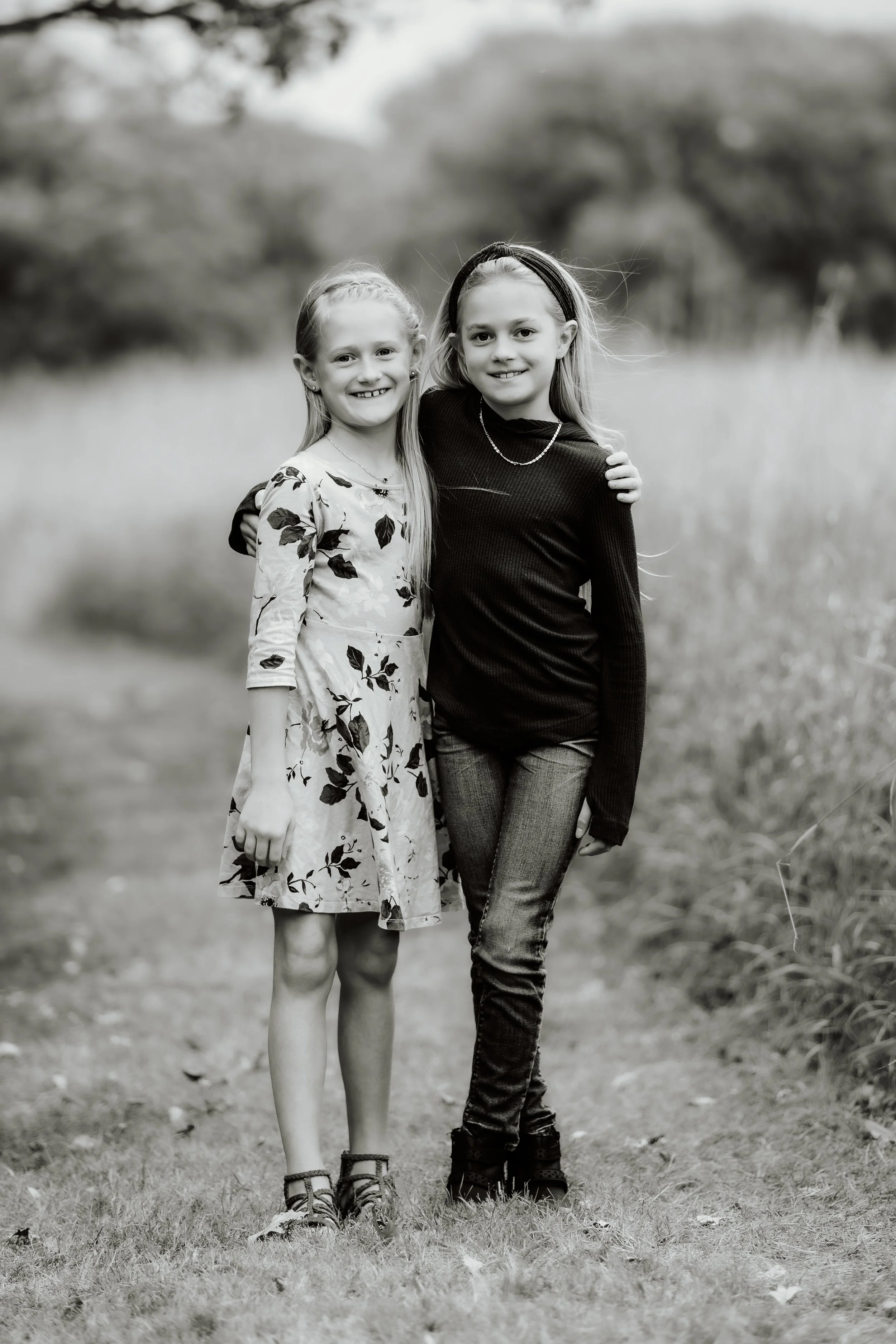 Two young girls standing close together outdoors, smiling at the camera, in a natural setting with blurred trees in the background in Sioux Falls, South Dakota at Good Earth State Park.