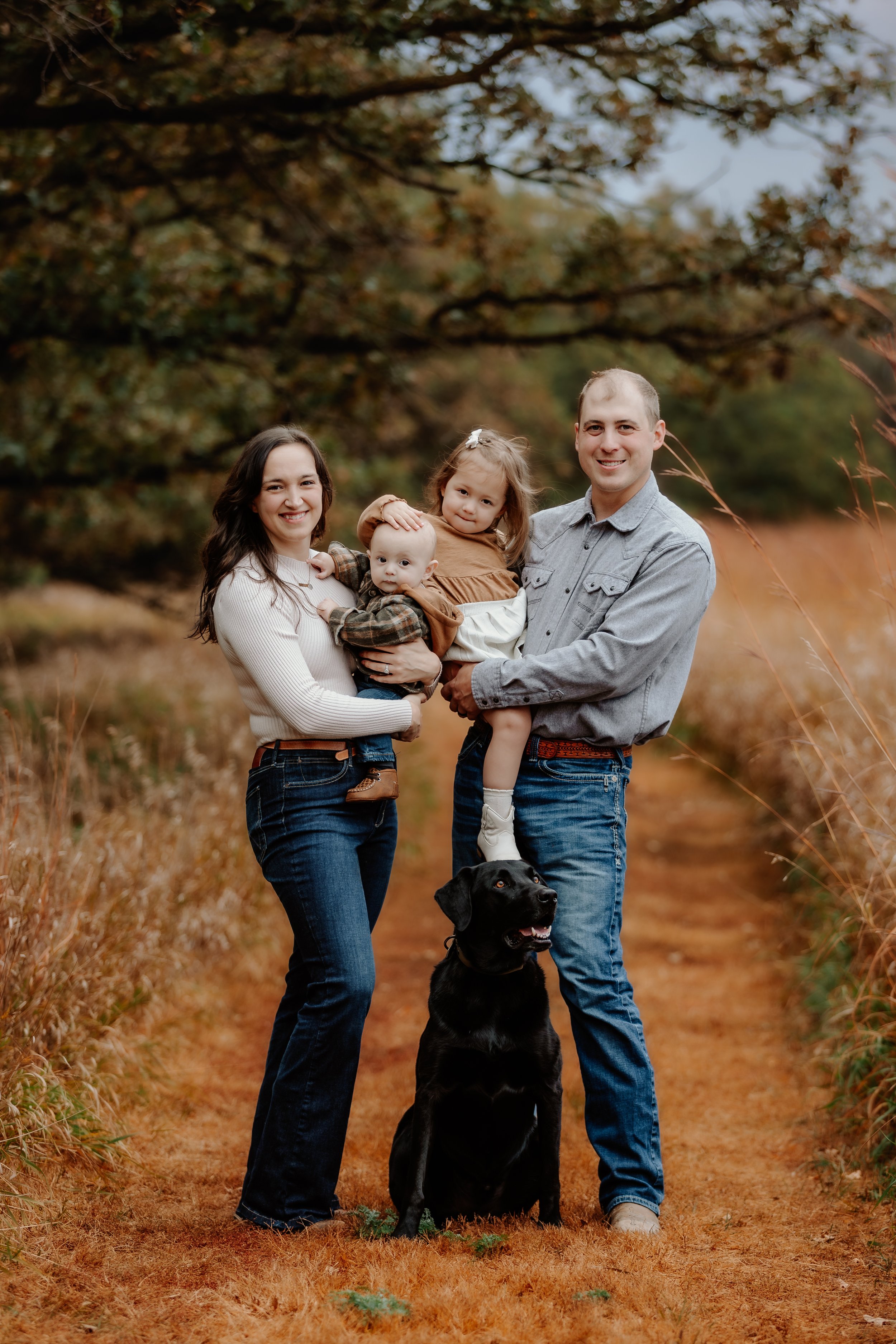 Family of four with dog outdoors during fall, standing on a dirt path with trees and autumn foliage in the background in Sioux Falls, South Dakota at Good Earth State Park.
