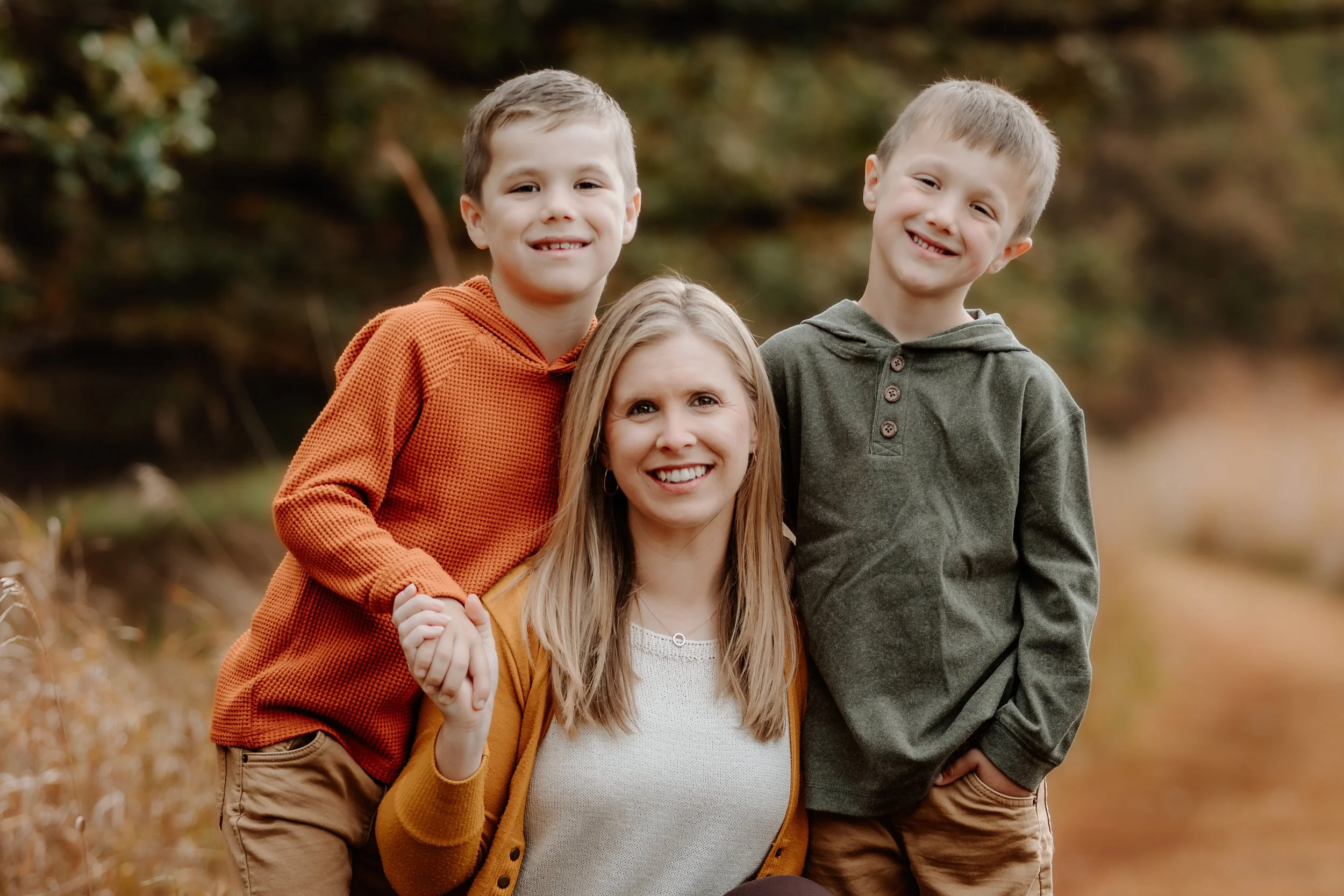 A woman with two young boys outdoors on a fall day, holding hands and smiling at the camera in Sioux Falls, South Dakota at Good Earth State Park.