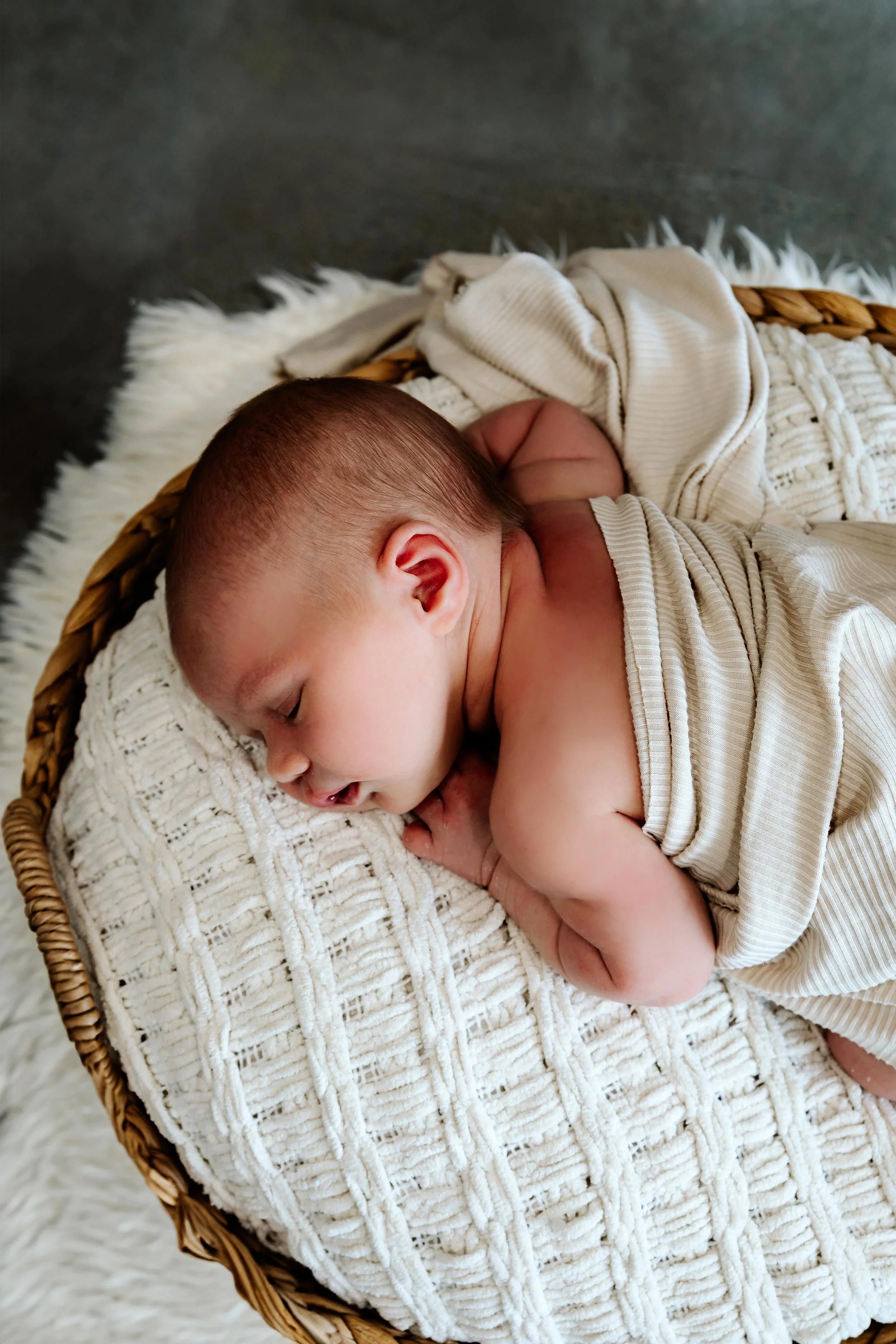 A sleeping baby lying on a soft, cream-colored knitted blanket in a wicker basket at The White Space Studio in Sioux Falls, South Dakota.