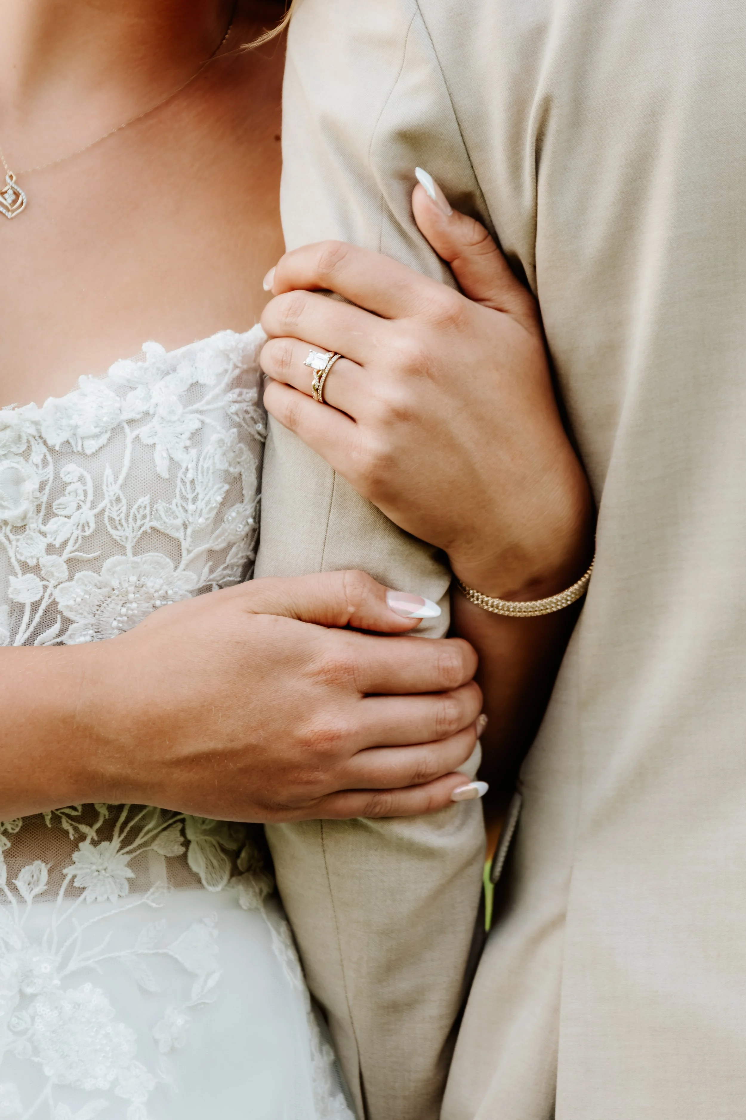 Close-up of a woman wearing a wedding dress and a man wearing a beige suit embracing each other, showing wedding rings and jewelry in Luverne, MN.