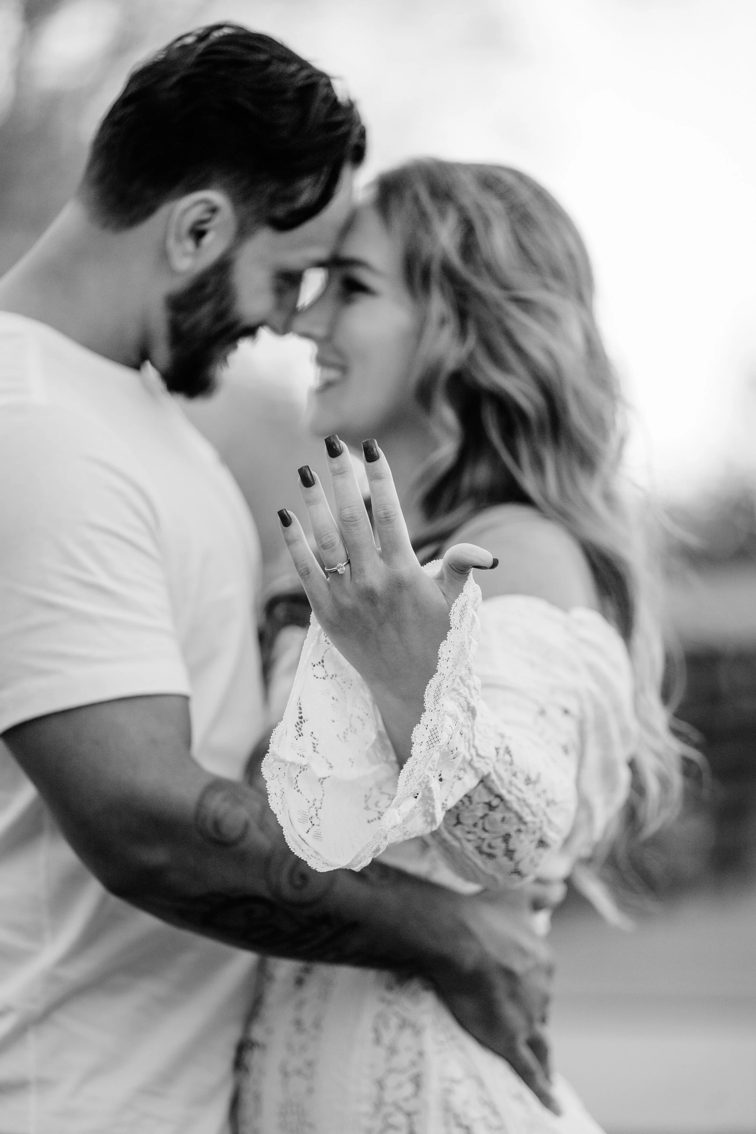 A black and white photo of a couple intimately close, smiling, with their foreheads touching, holding each other. The woman's hand is visible, showing an engagement ring at Mary Jo Wegner Arboretum in Sioux Falls, South Dakota.