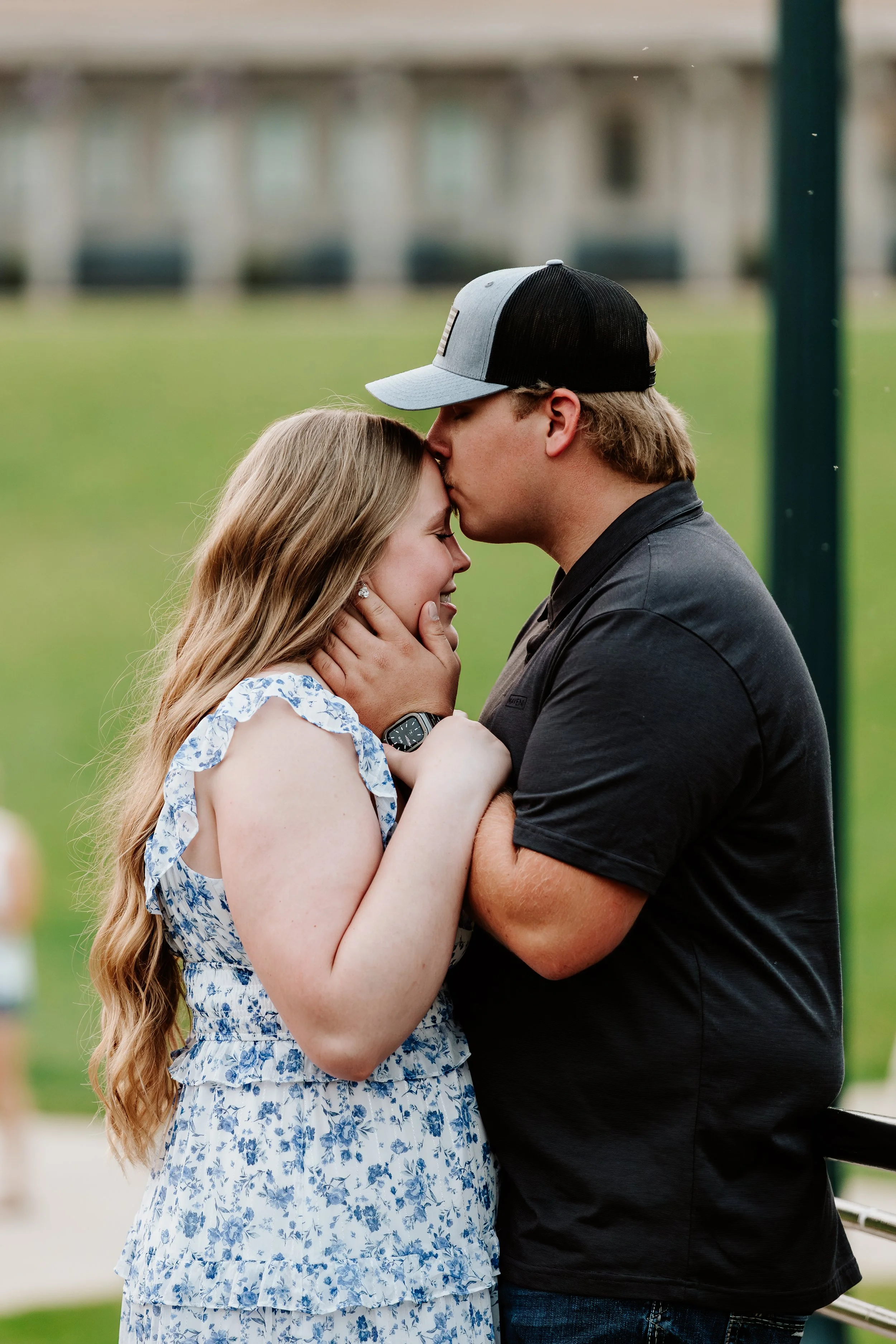 A man kissing a woman on the forehead while holding her face, in an outdoor setting with a grassy background and a building in the distance.
