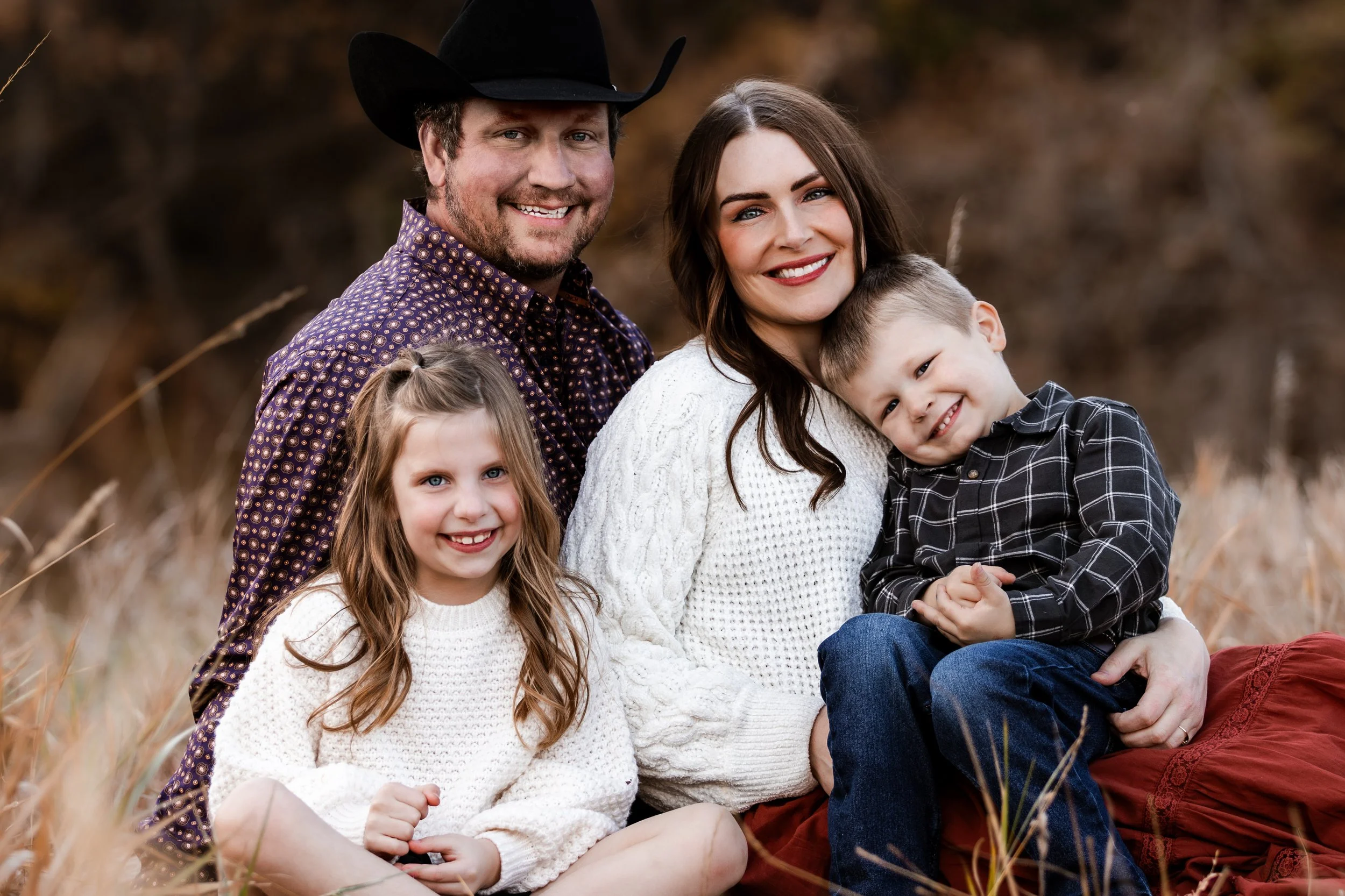 A happy family of four sitting outdoors in a field, smiling at the camera at Legacy Park in Sioux Falls, South Dakota.