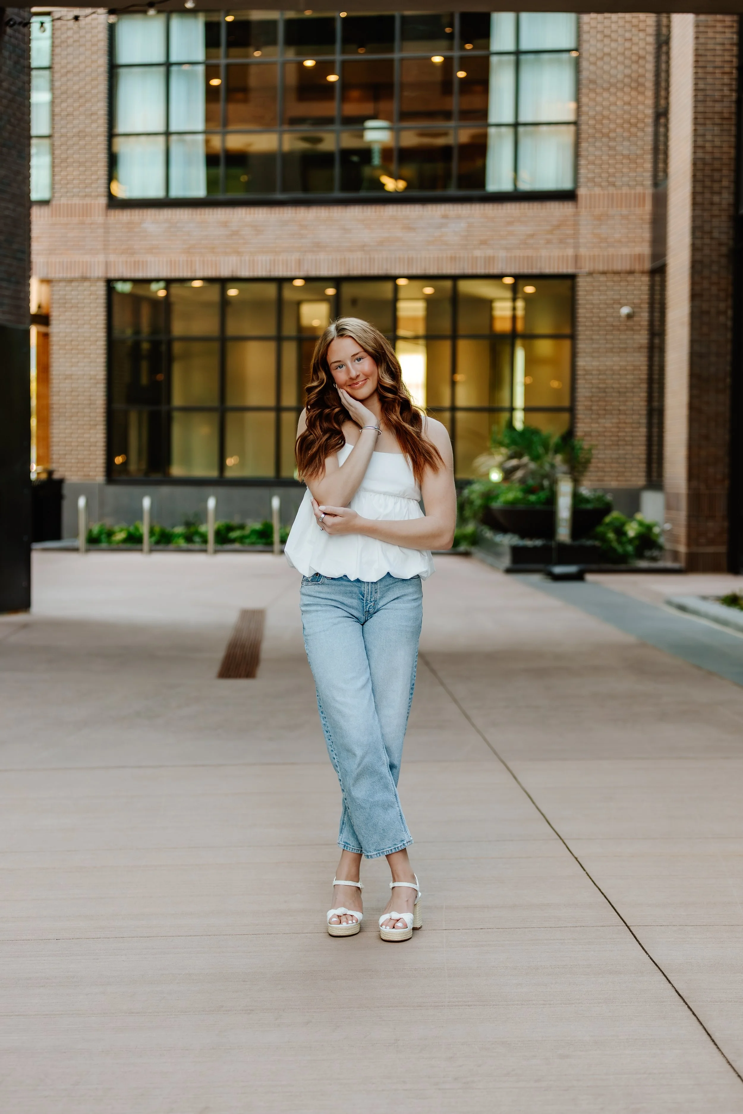 A young woman with long wavy brown hair, wearing a white sleeveless top, light blue jeans, and white platform sandals, standing outdoors in front of a modern brick building with large glass windows in Downtown Sioux Falls, South Dakota.