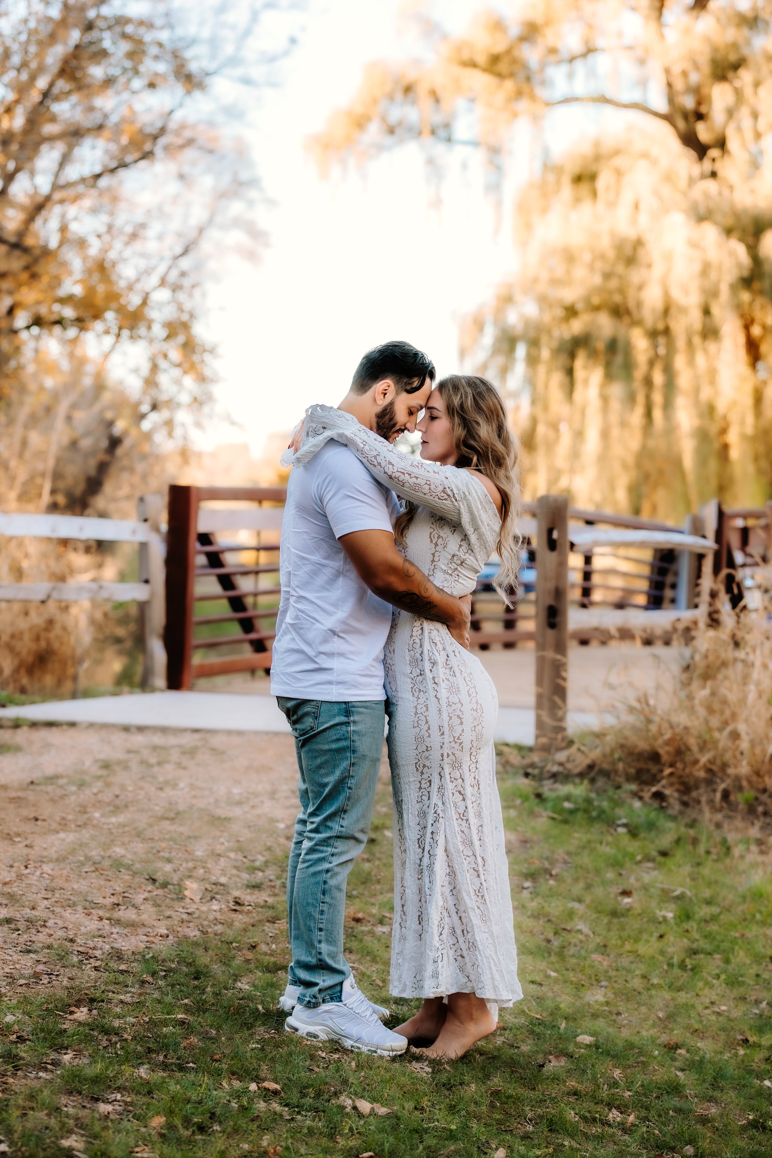 A couple embracing outdoors in a park during golden hour, with trees and a wooden bridge in the background at Mary Jo Wegner Arboretum in Sioux Falls, South Dakota.