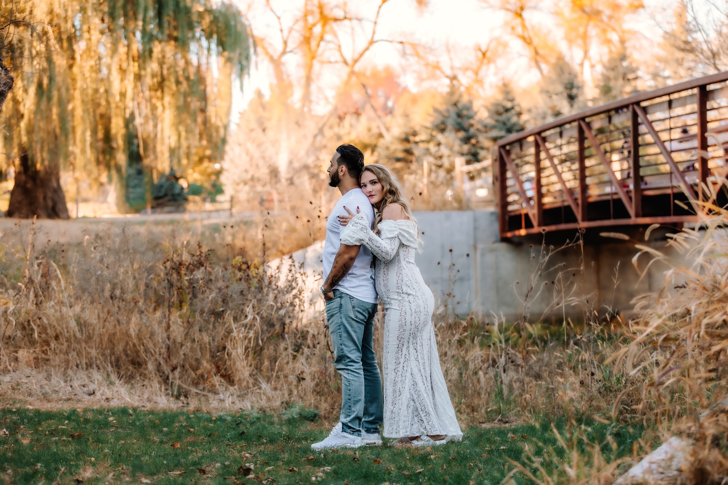 A couple standing close together outdoors during autumn, the woman leaning on the man's shoulder, with trees and a bridge in the background at Mary Jo Wegner Arboretum in Sioux Falls, South Dakota.
