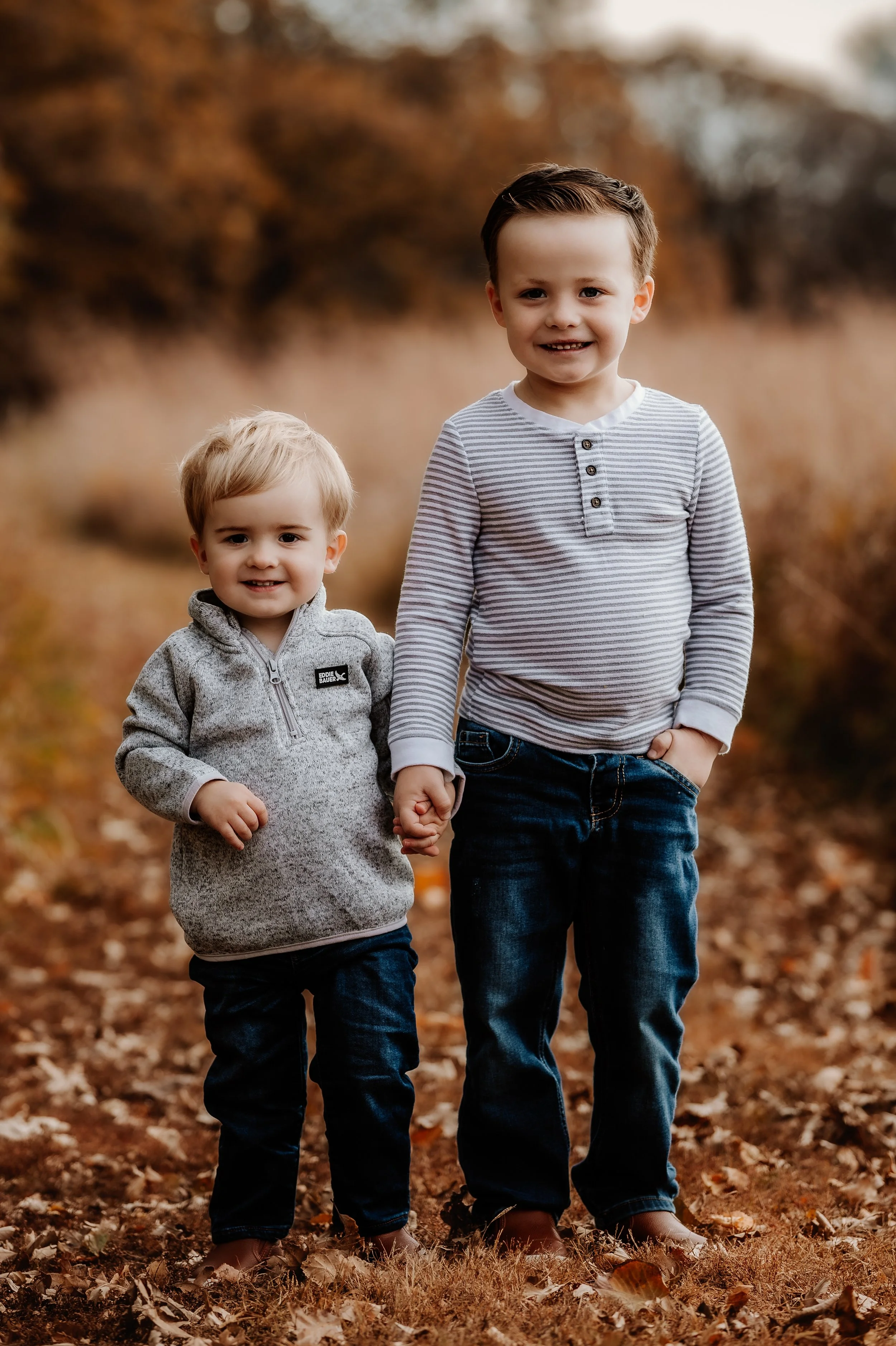 Two young boys walking outdoors on a fall day, holding hands, with fallen leaves on the ground and trees in the background in Sioux Falls, South Dakota at Good Earth State Park.