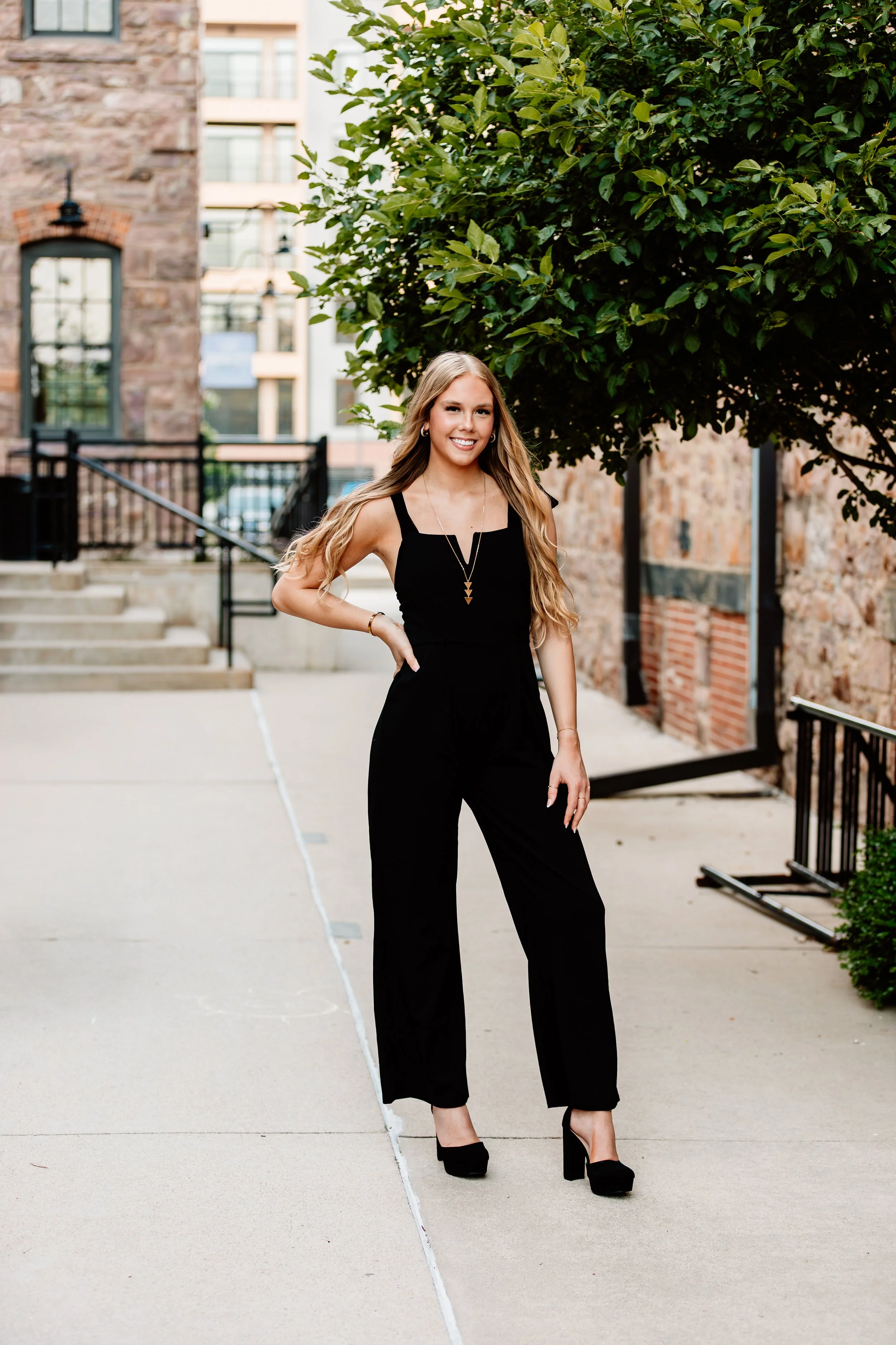 A smiling young woman in a black jumpsuit and high heels standing outdoors on a sidewalk with green trees and brick buildings in the background in Downtown Sioux Falls, South Dakota.