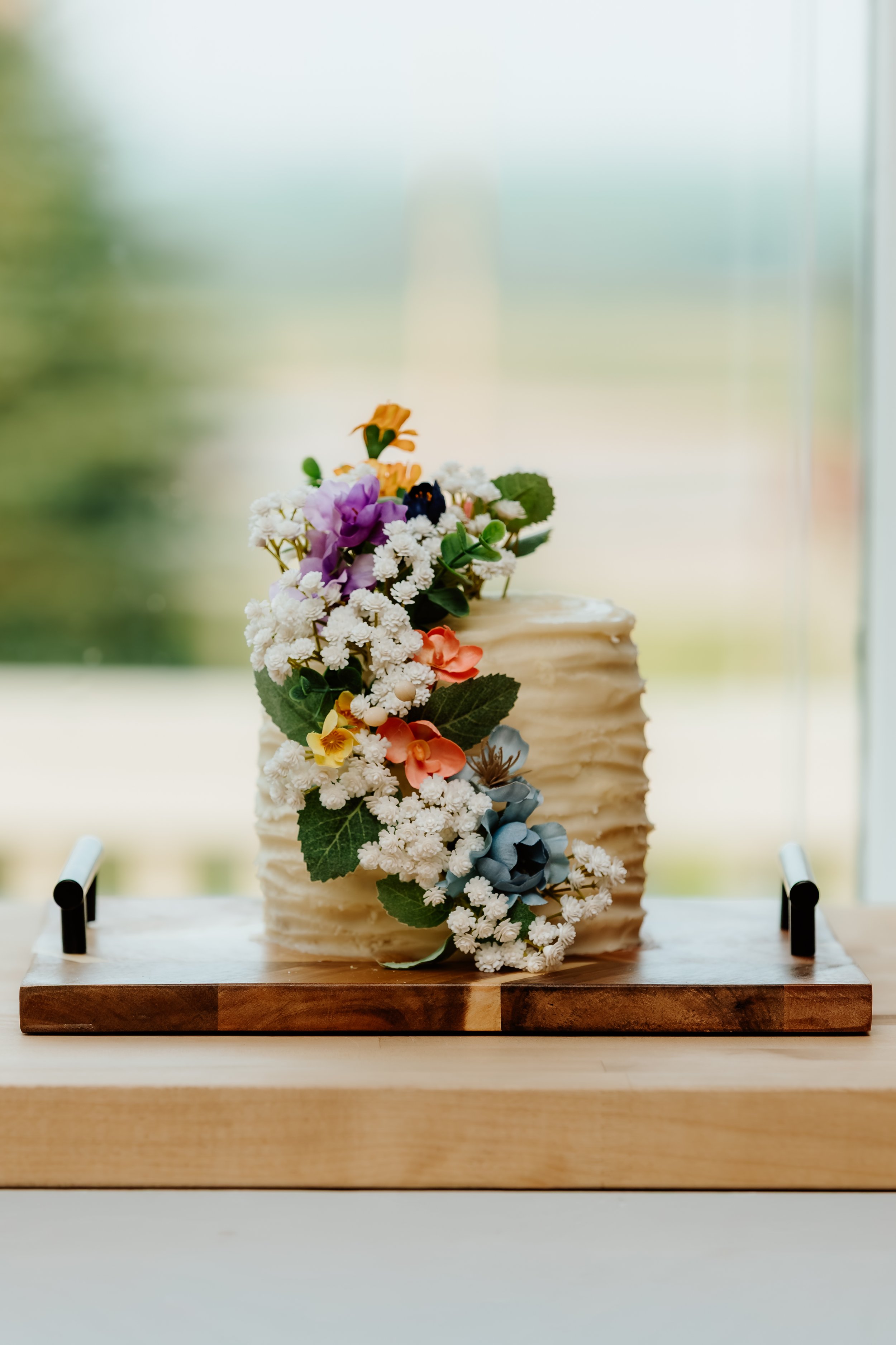 A decorative cake with white textured frosting, topped with colorful artificial flowers and green leaves, placed on a wooden board in Luverne, MN.
