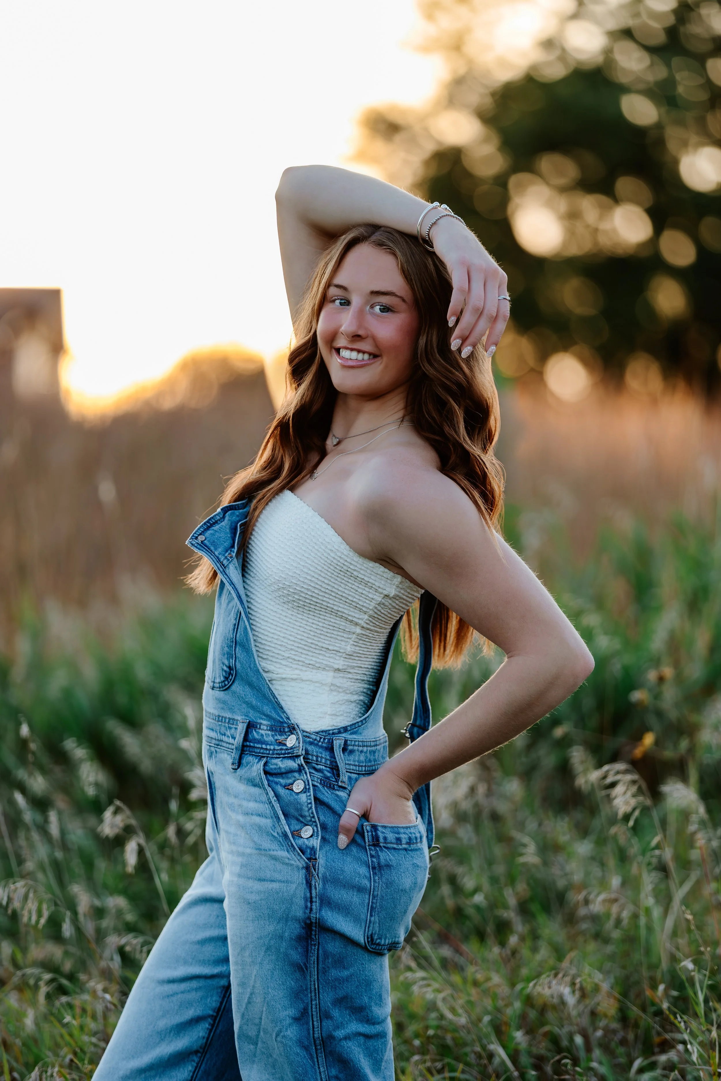 A young woman with long, wavy red hair standing outdoors in a field at sunset, wearing a white strapless top and blue denim overalls, smiling and posing with one hand behind her head at Good Earth State Park in Sioux Falls, South Dakota.
