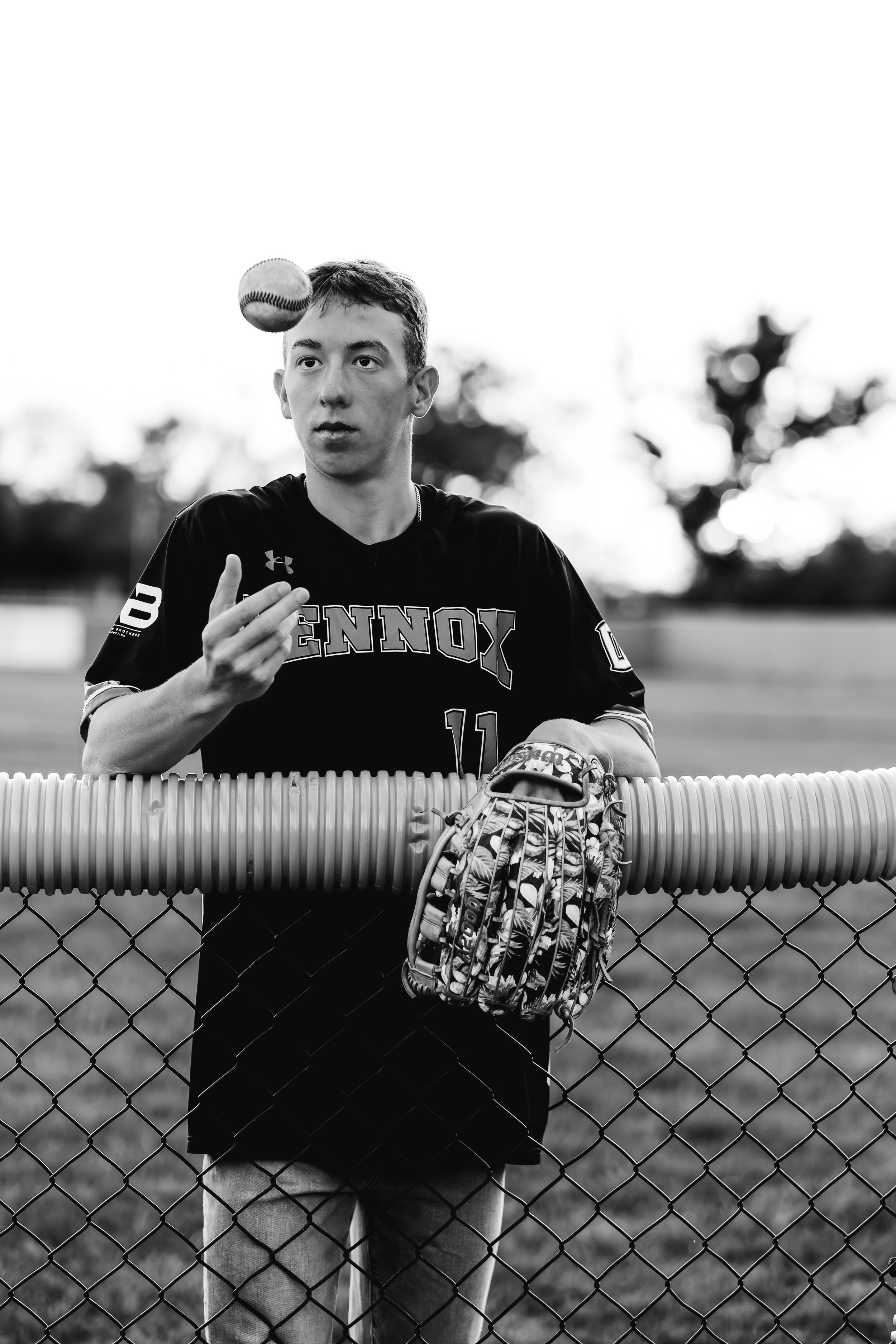A young man in a sports jersey stands behind a chain-link fence, holding a baseball glove with a baseball spinning in the air above his head, outdoors during daytime in Lennox, South Dakota.