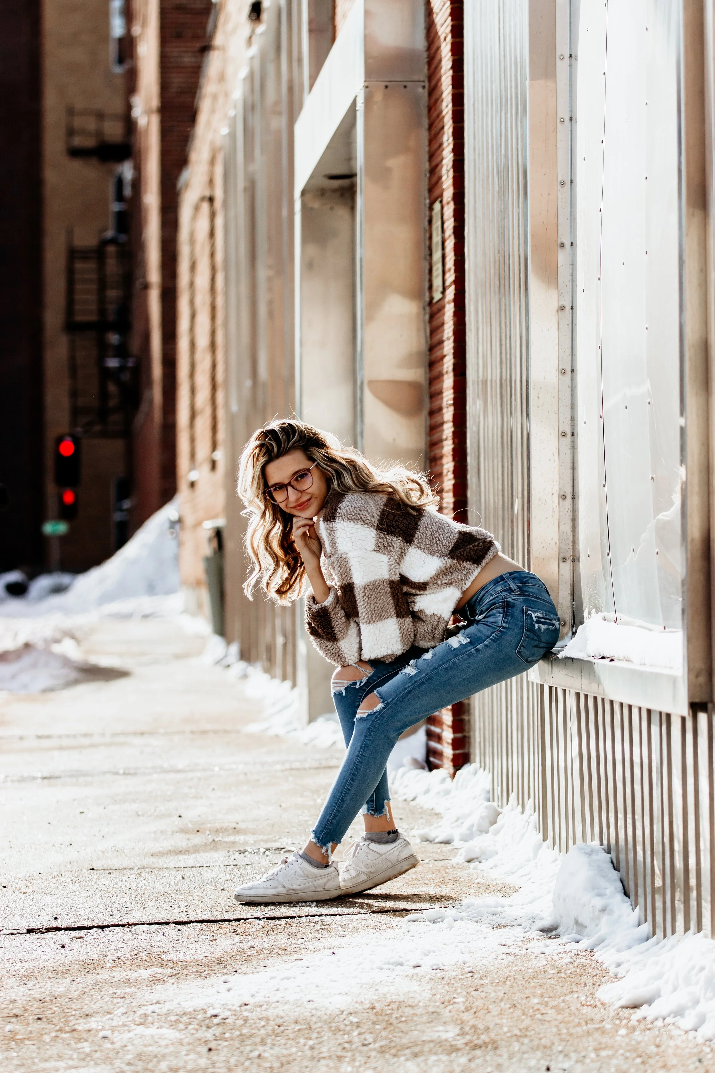 A young woman with blonde wavy hair, glasses, and a checkered sweater crouching on a snowy sidewalk next to a metal and brick building during winter in Downtown Sioux Falls, South Dakota.