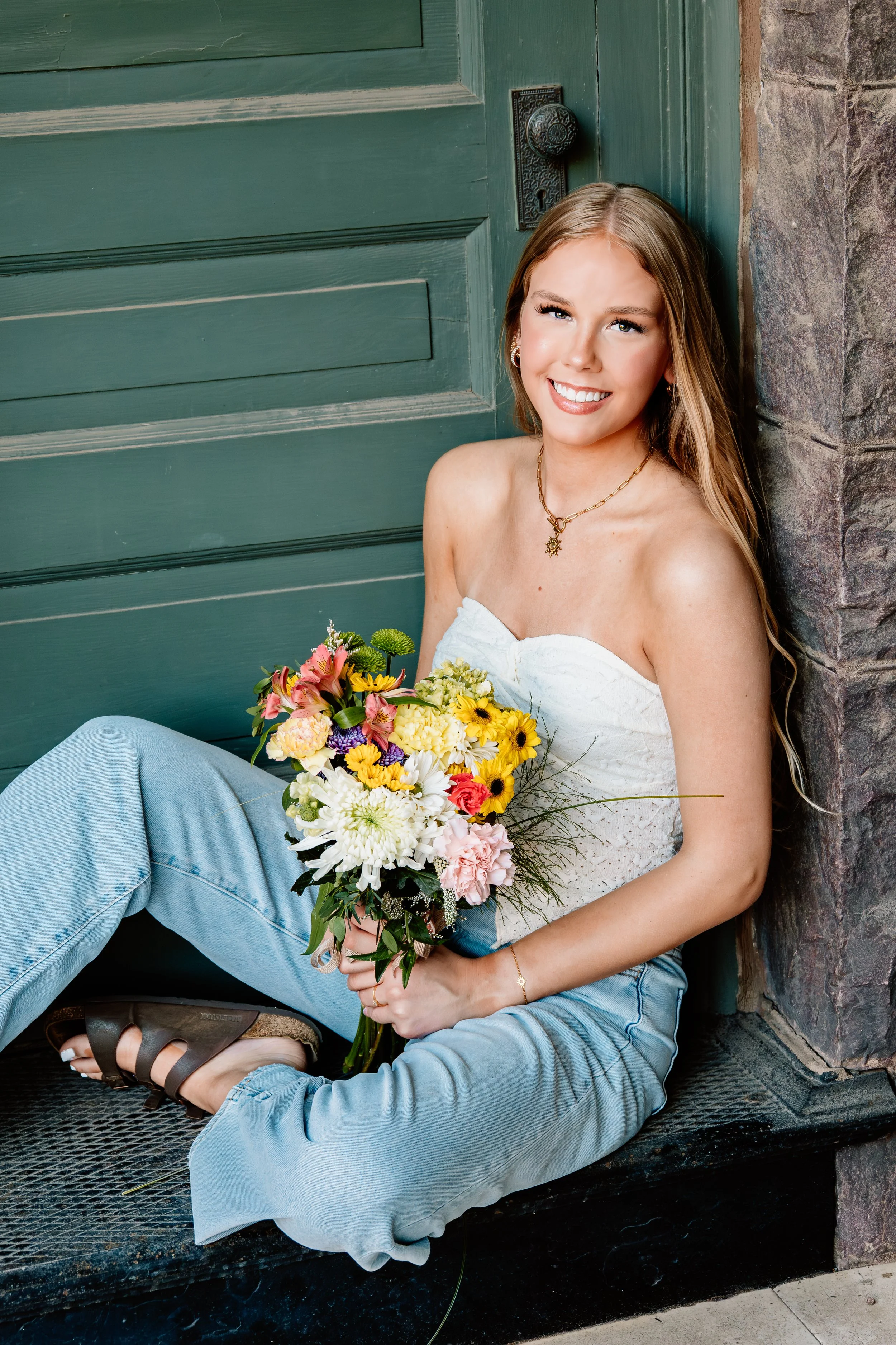 Young woman with long blonde hair, sitting on the ground outdoors and leaning against a stone wall with a green door behind her. She is smiling, wearing a white strapless top, light blue jeans, and brown sandals in Downtown Sioux Falls, South Dakota.