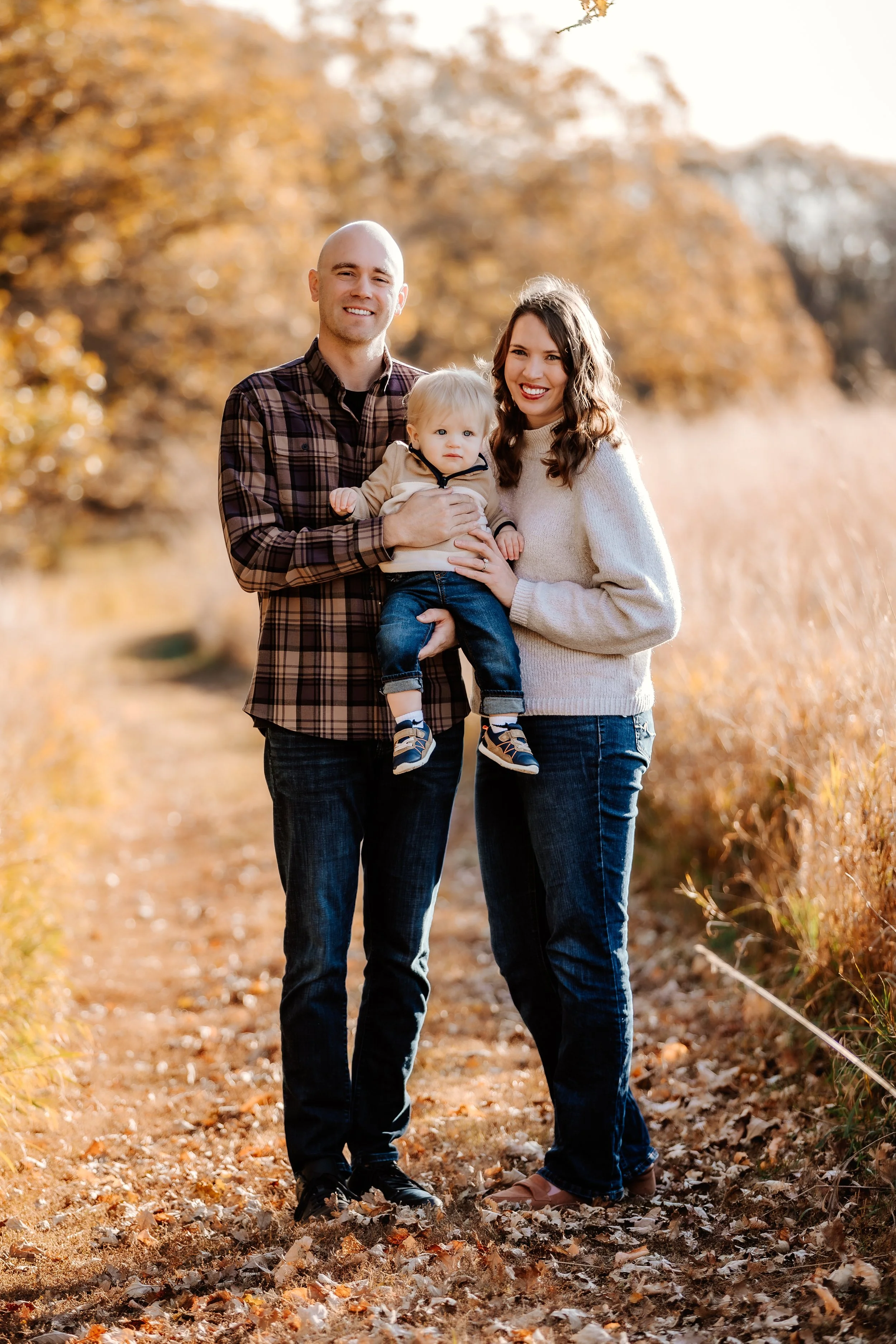 A family of three standing outdoors on a fall day, with golden leaves and trees in the background. The father is holding a young child, and the mother is standing beside them. They are smiling in Sioux Falls, South Dakota at Good Earth State Park.