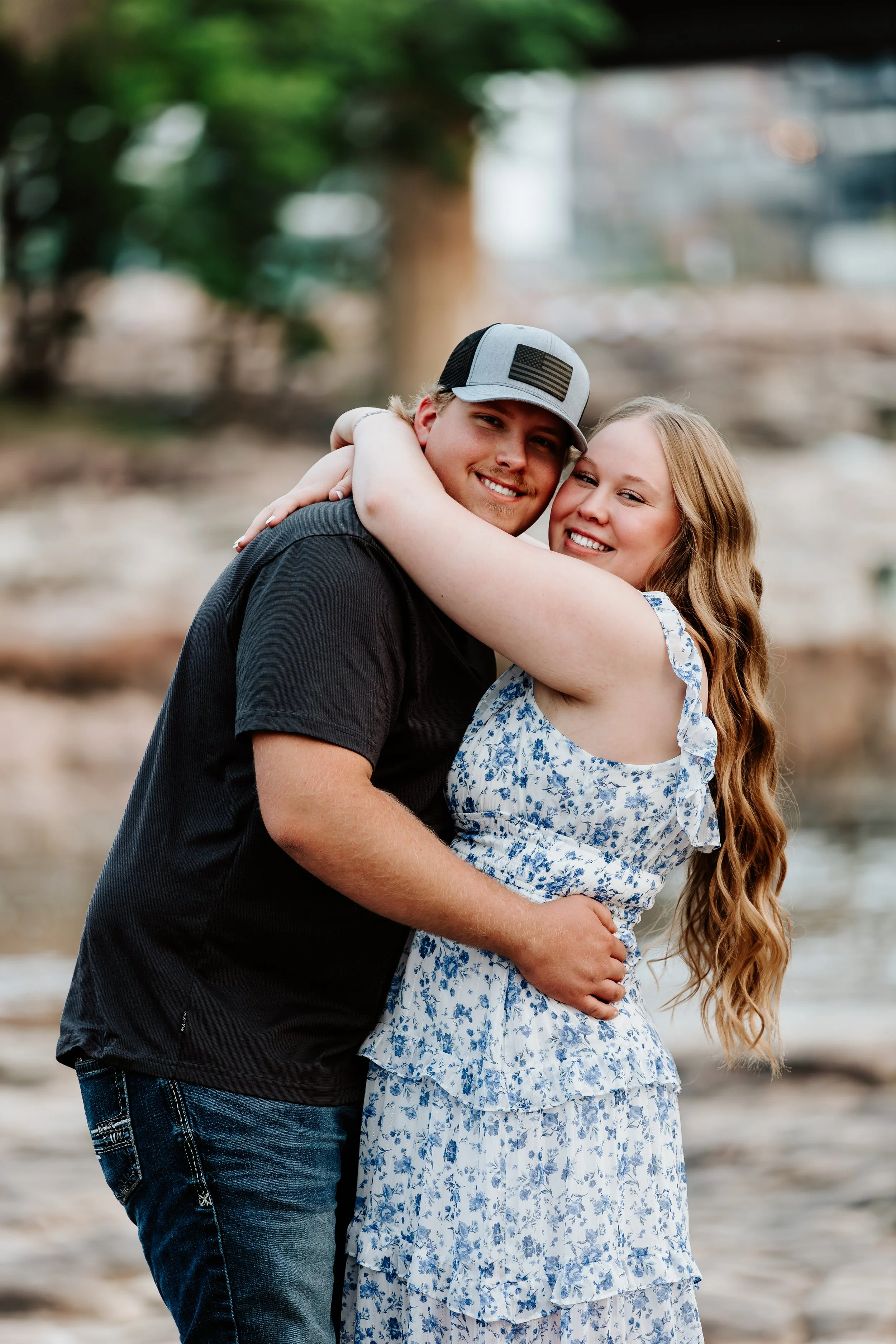 A smiling young man and woman hugging outdoors near a body of water, with trees and rocks in the background at The Falls Park in Sioux Falls, South Dakota.