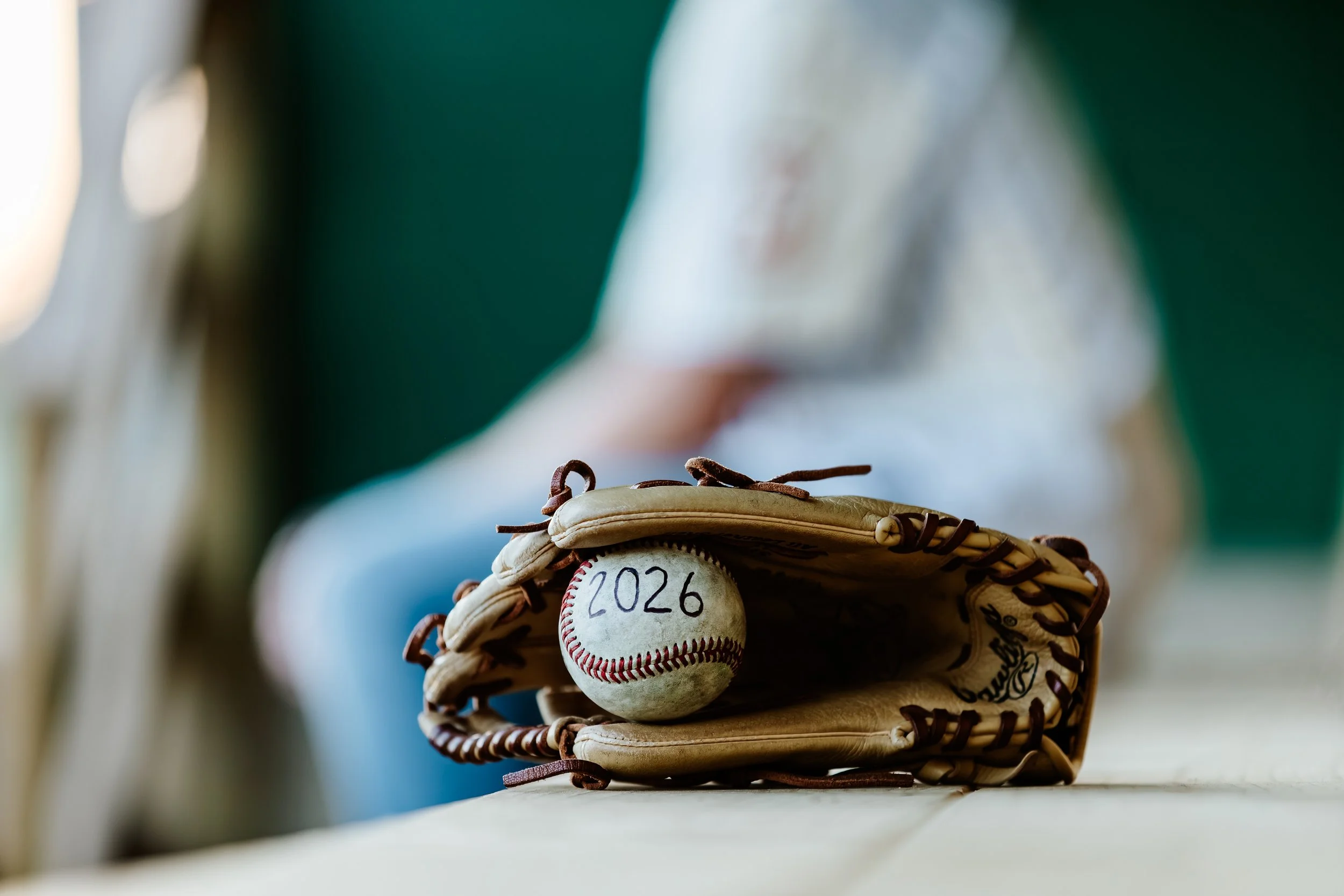 A baseball glove holding a baseball with '2026' written on it, resting on a wooden surface.