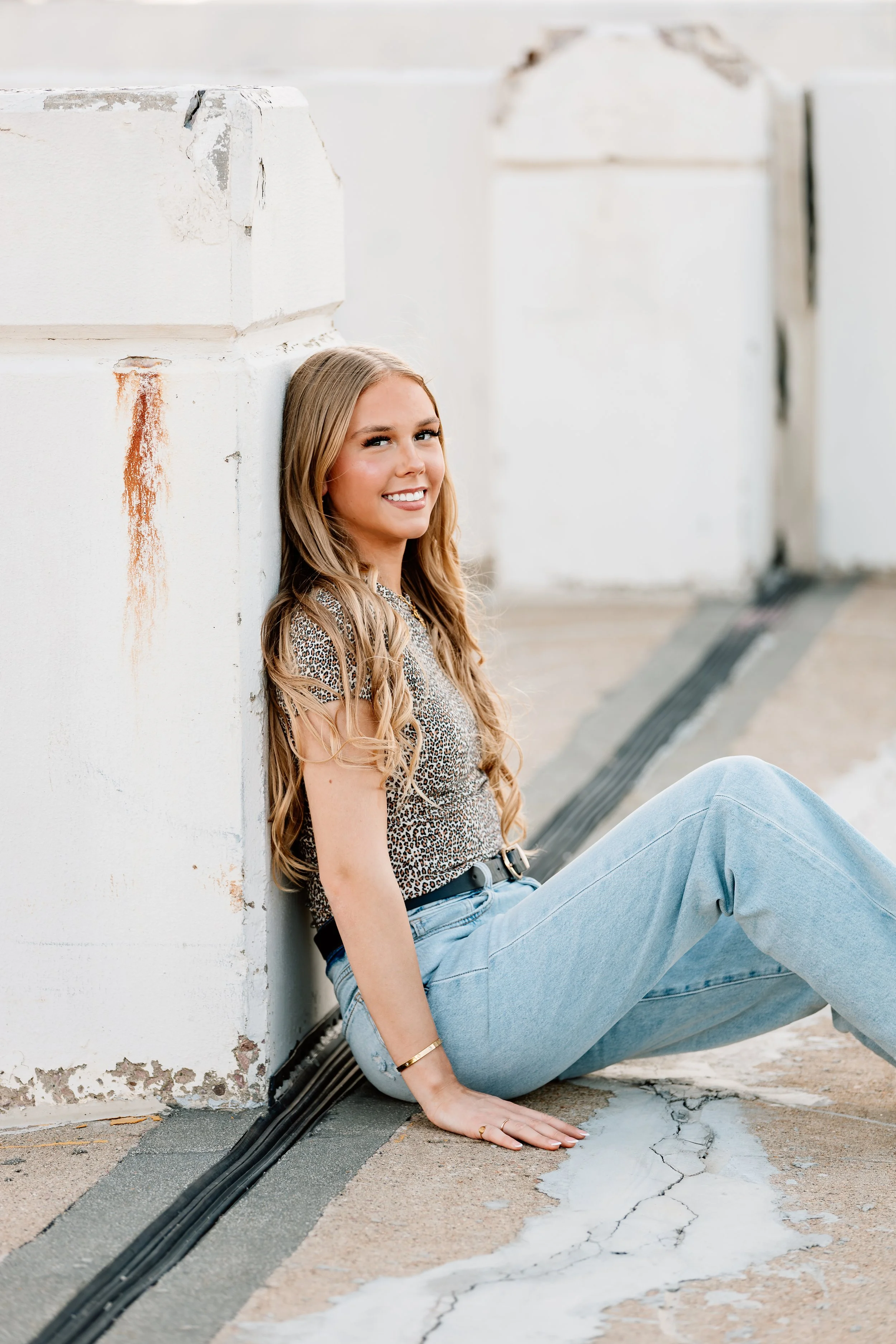 A young woman with long blonde hair, wearing a leopard print top and light blue jeans, is sitting on the ground, leaning against a white concrete wall with rust stains, smiles at the camera on a rooftop or urban setting in Downtown Sioux Falls, South