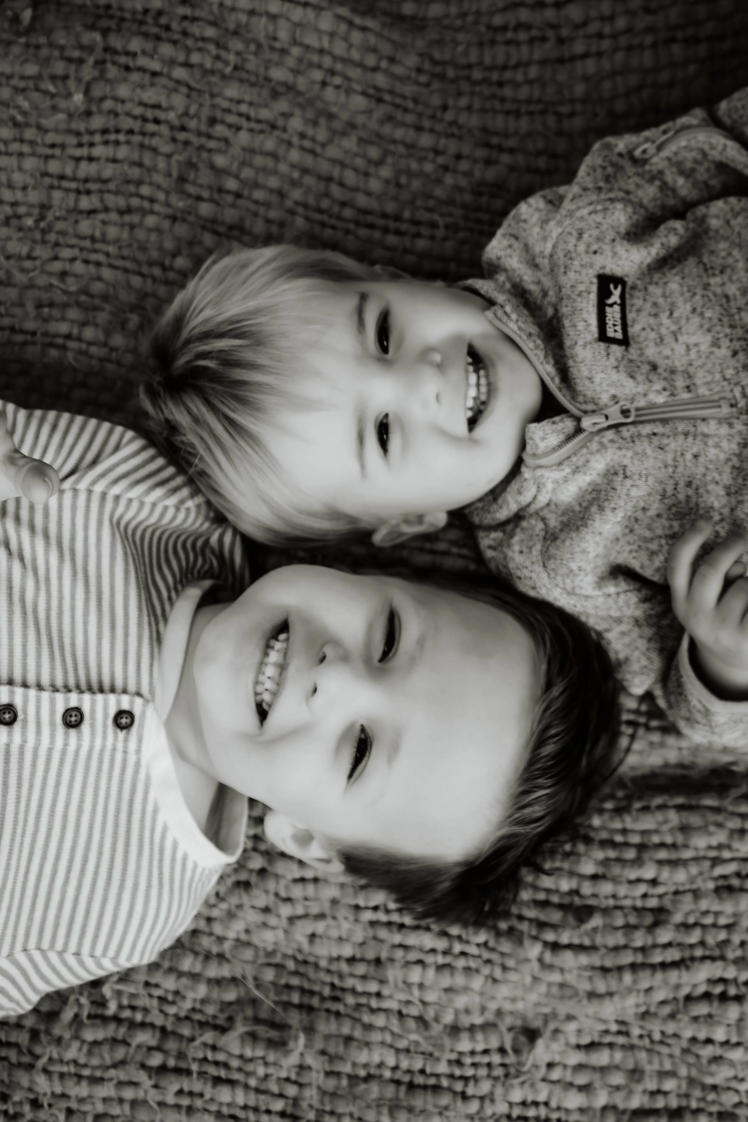 Two young children, one with light-colored hair and one with dark-colored hair, lying on a textured carpet, smiling and looking up at the camera in a black and white photo in Sioux Falls, South Dakota at Good Earth State Park.