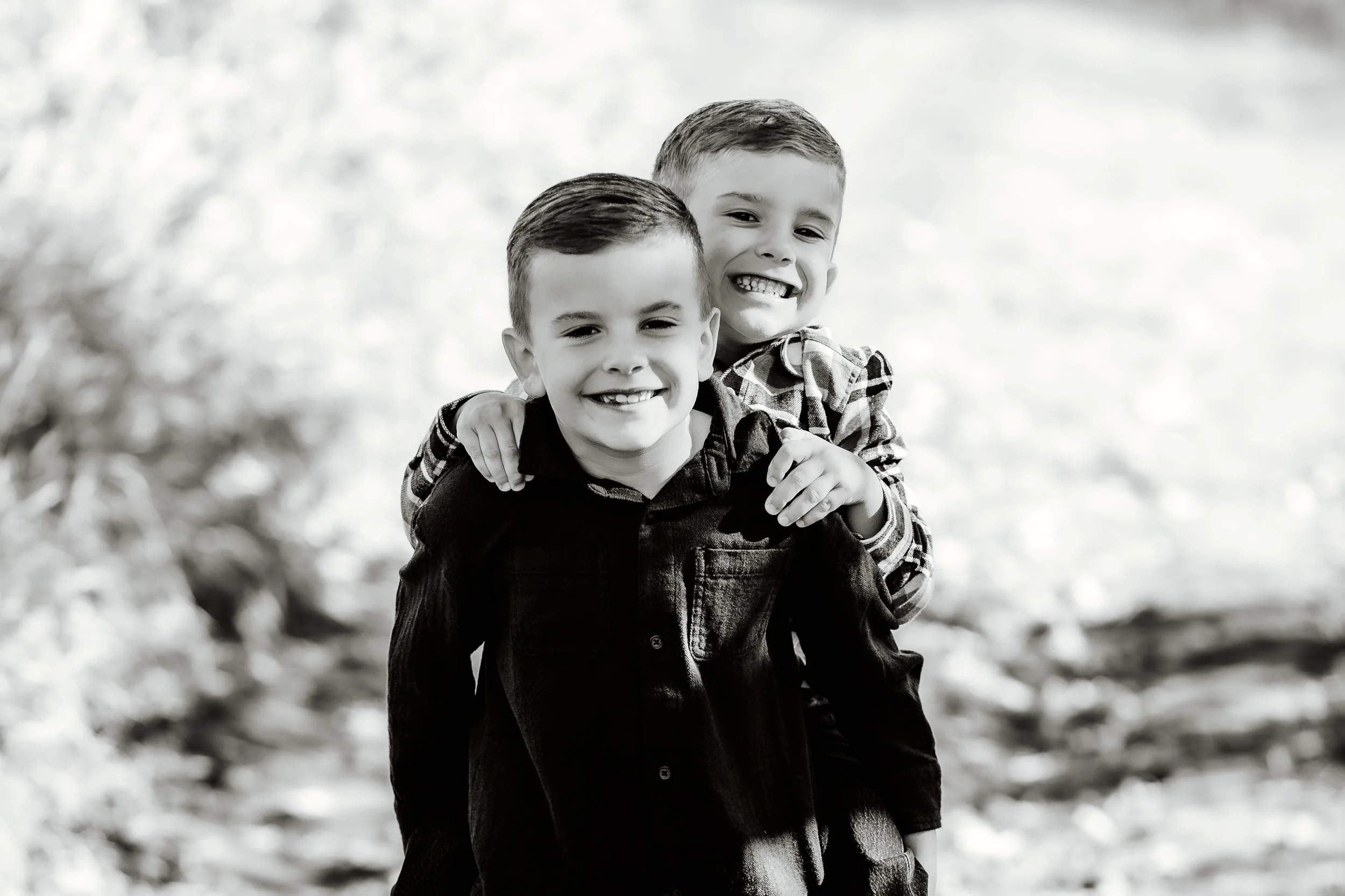 Two young boys smiling, with one giving the other a piggyback ride, outdoors surrounded by blurred natural background in Sioux Falls, South Dakota at Good Earth State Park.