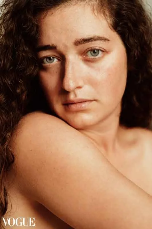 Portrait serré d'une jeune femme blanche aux yeux bleus, qui regarde frontalement l'objectif. Ses cheveux longs bouclés forment un cadre sur les côtés de l'image.