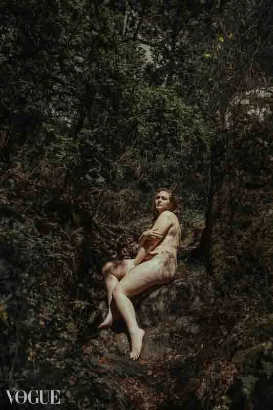 Photographie d'une femme ronde assise sur un rocher dans un chemin de forêt. Elle a l'air détendue et sereine, et regarde l'objectif de la photographe.