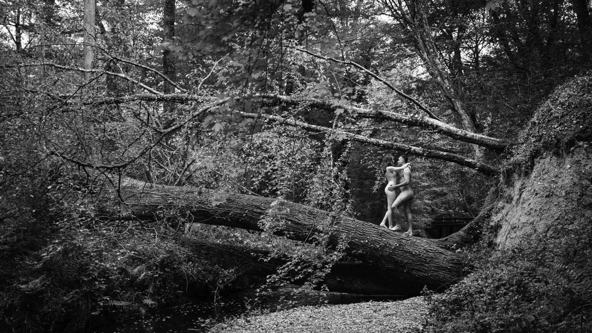 Photo noir et blanc d'un couple en forêt. Le couple est nu et s'enlace, debout sur un tronc d'arbre tombé au dessus d'une rivière.