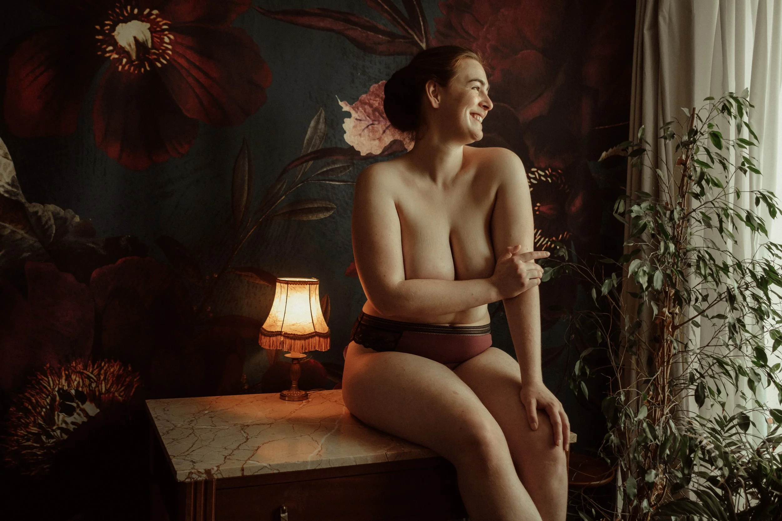 Une femme souriante portant juste une culotte de coton bordeaux est assise sur une table, entre une lampe et une plante, devant un fond fleuri romantique.