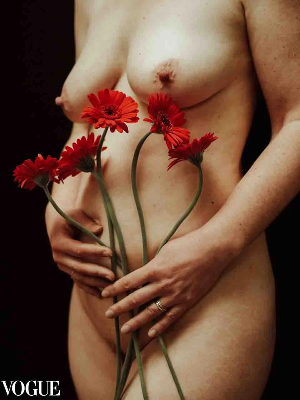 Photographie en plan serré sur le corps d'une femme blanche nue. Devant son ventre, elle tient délicatement des fleurs gerberas rouges.
