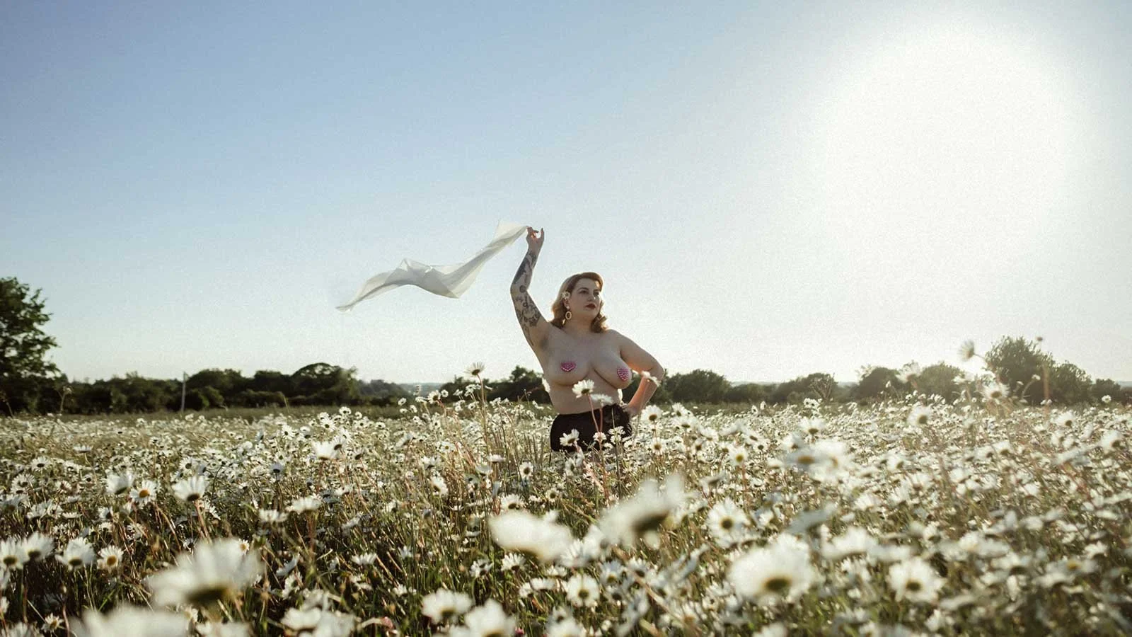 Photographie d'une femme blonde pulpeuse portant des cache-tétons en forme de coeurs, se tenant debout dans un champ de marguerites. Elle tient au dessus d'elle un foulard en soie blanche qui vole au vent.