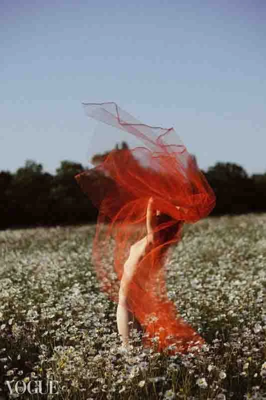 Photographie d'une femme nue dans un champ de marguerites, qui lance un grand voile rouge au dessus d'elle, qui forme comme une flamme autour d'elle.
