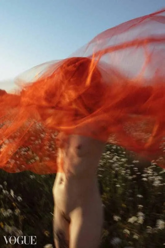Photographie en flou artistique d'une femme nue dans un champ de marguerites. Elle fait tourner un voile de tulle rouge autour d'elle.