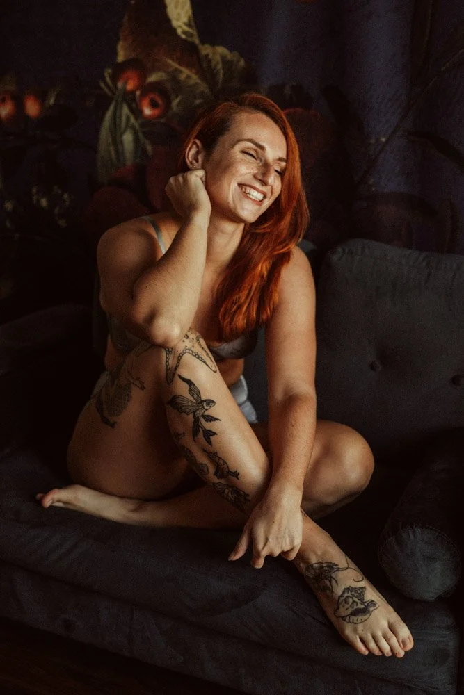 Photo d'une jeune femme rousse en lingerie grise avec un grand sourire et assise sur un canapé.