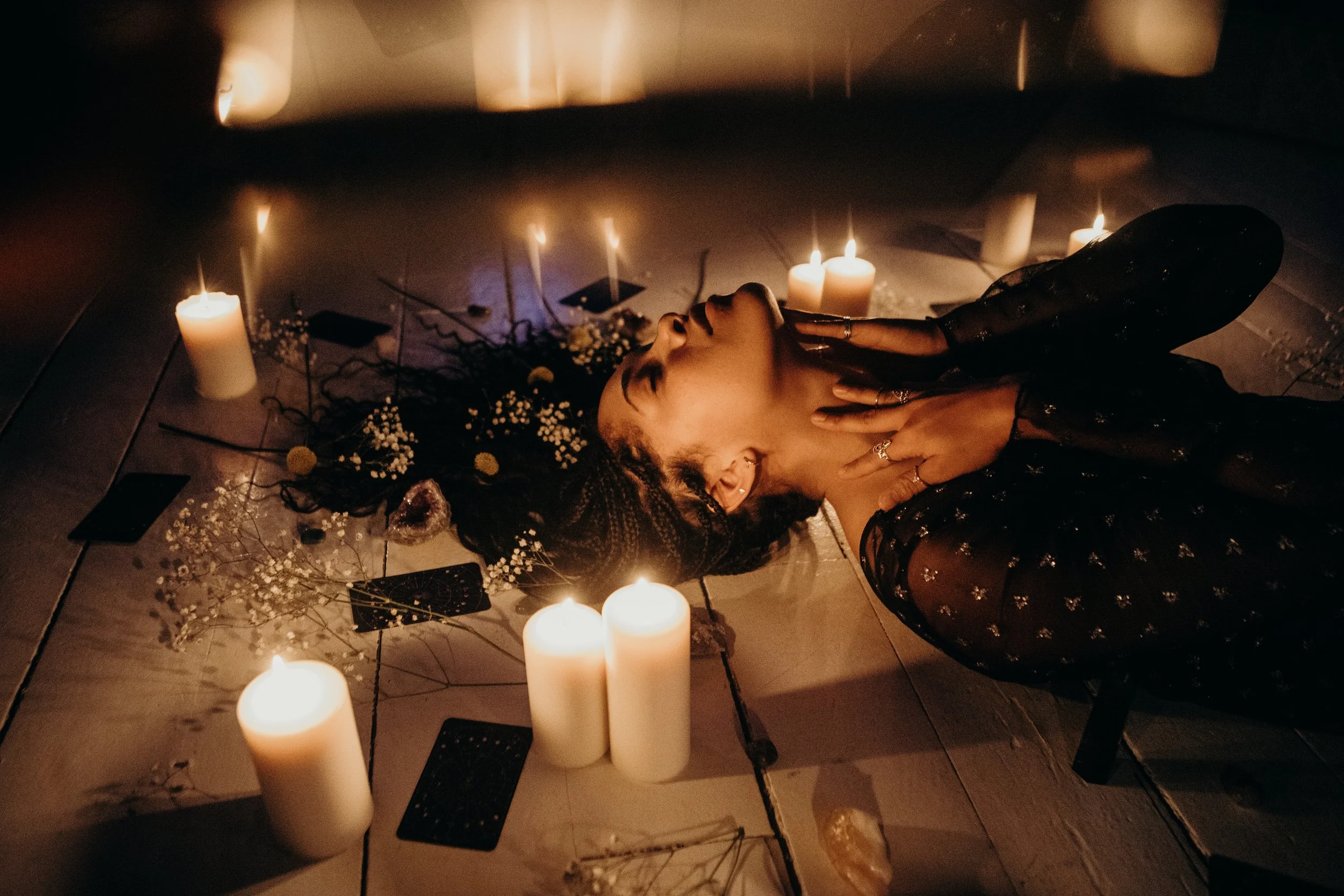 Gabriela Nova with braided hair lying on the floor surrounded by lit candles, tarot cards, and flowers, with her eyes closed, in a dimly lit setting.