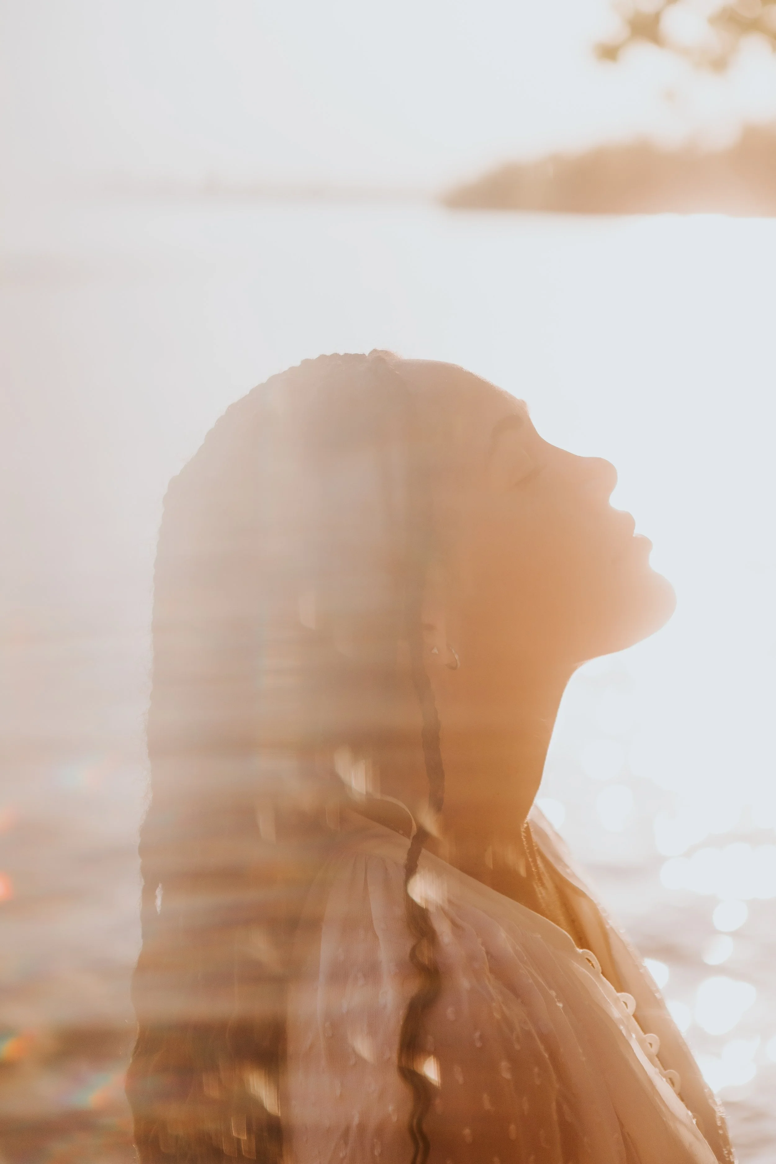 Gabriela Nova enjoys a moment with her eyes closed, basking in sunlight near water with her profile visible, and long hair flowing with gentle waves.