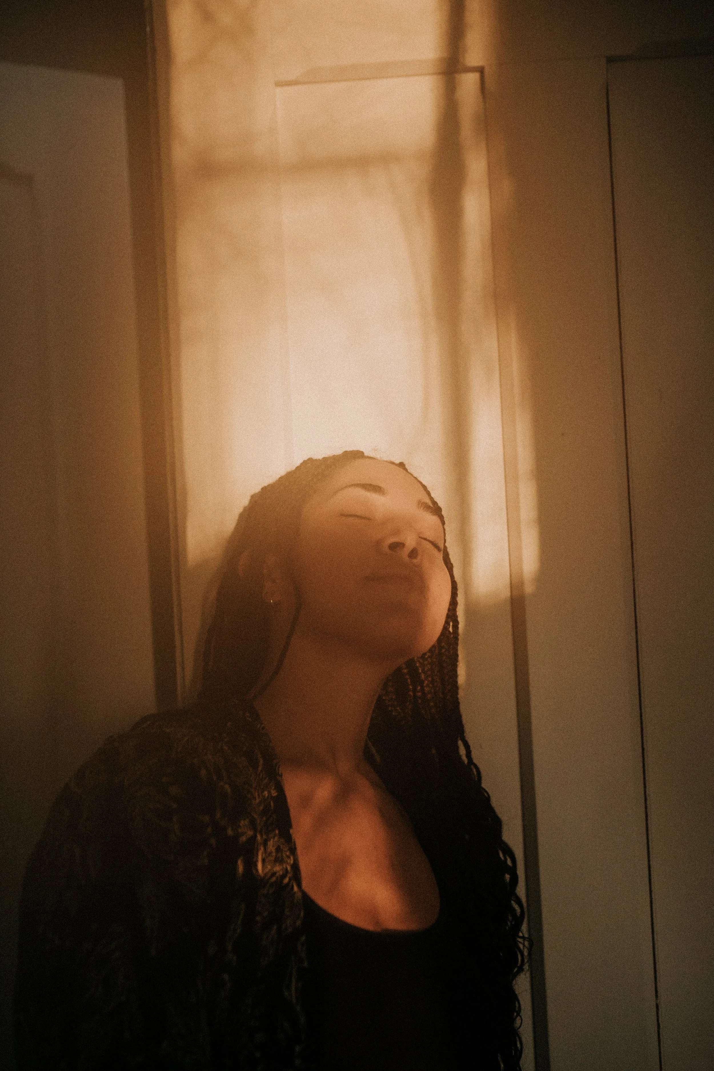 Gabriela Nova with braided hair leaning against a wall with eyes closed, illuminated by warm light.