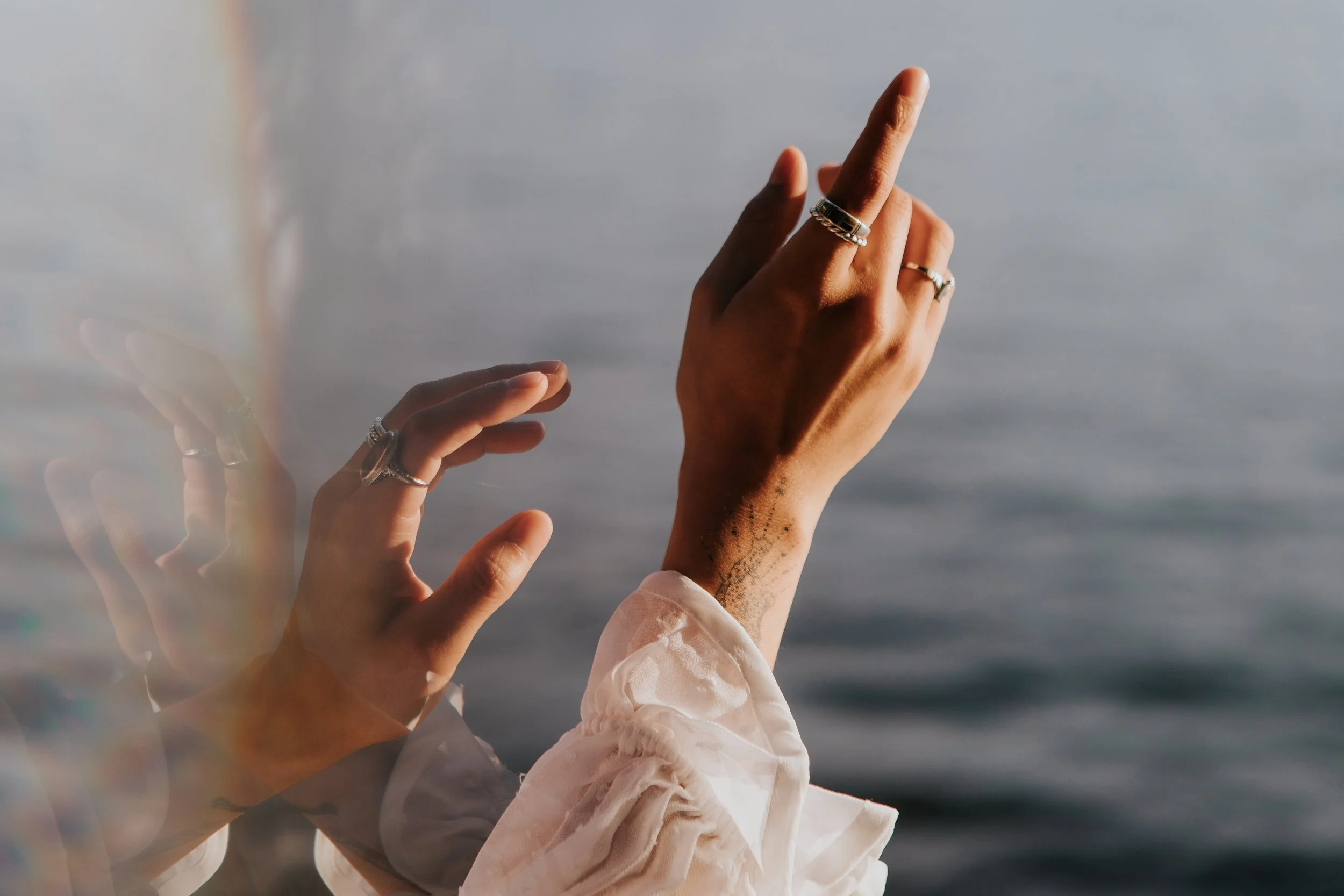 Two hands with rings, one raised with middle finger up near a body of water, the other hand making a pinching gesture. The person is wearing a white, ruffled sleeve.