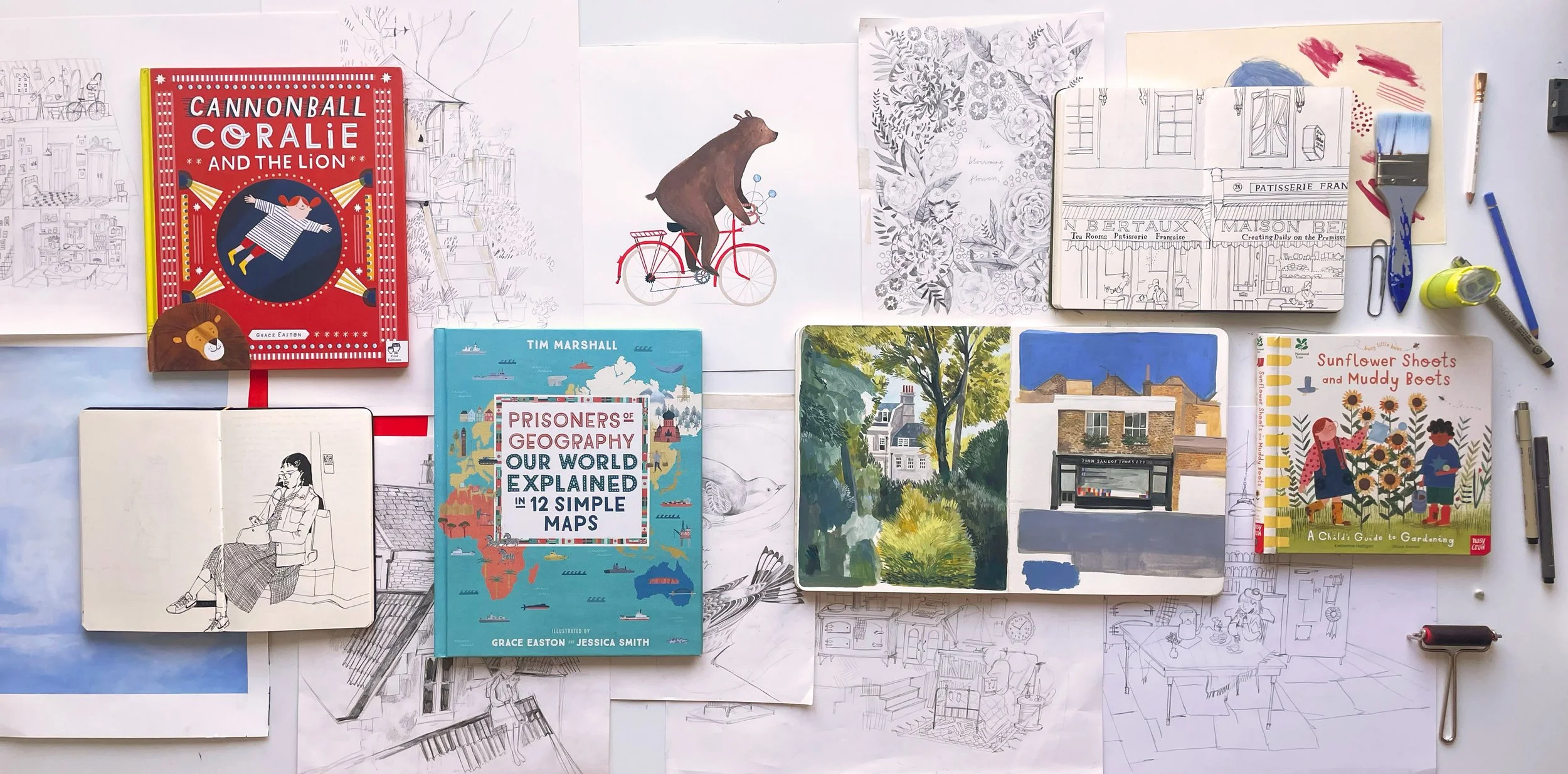 An artist's workspace with various sketches, booklets, and painting supplies arranged on a table. Visible items include illustrated books, architectural sketches, and art tools.