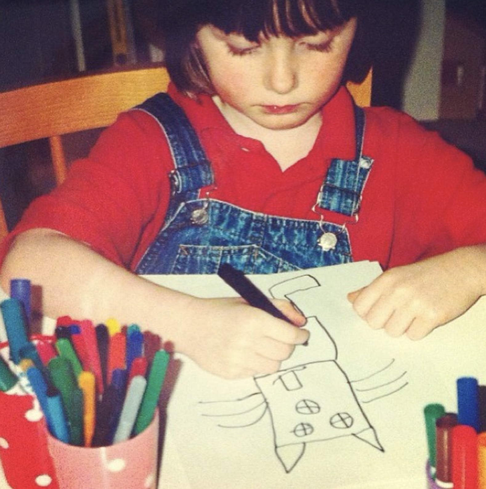 A young boy with dark hair wearing a red shirt and denim overalls, sitting at a table, coloring a drawing of a house and a rocket ship with markers.